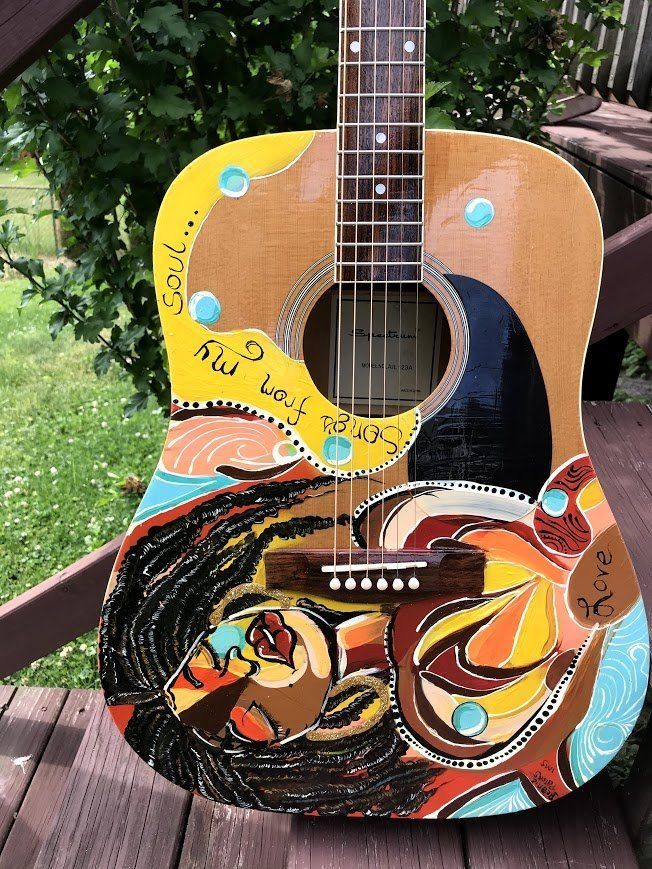 Colorful painted acoustic guitar with a face and swirling designs, outdoors against green foliage.