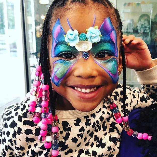 Smiling child with colorful butterfly face paint and pink bead braids, wearing a black-and-white spotted top