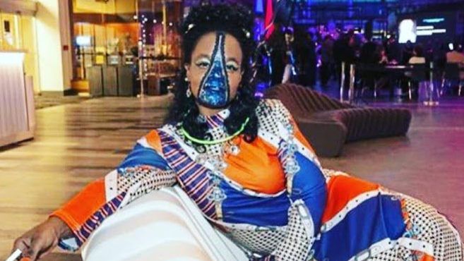 Person seated on a lounge chair in a colorful patterned outfit and face paint, in a dimly lit indoor lounge