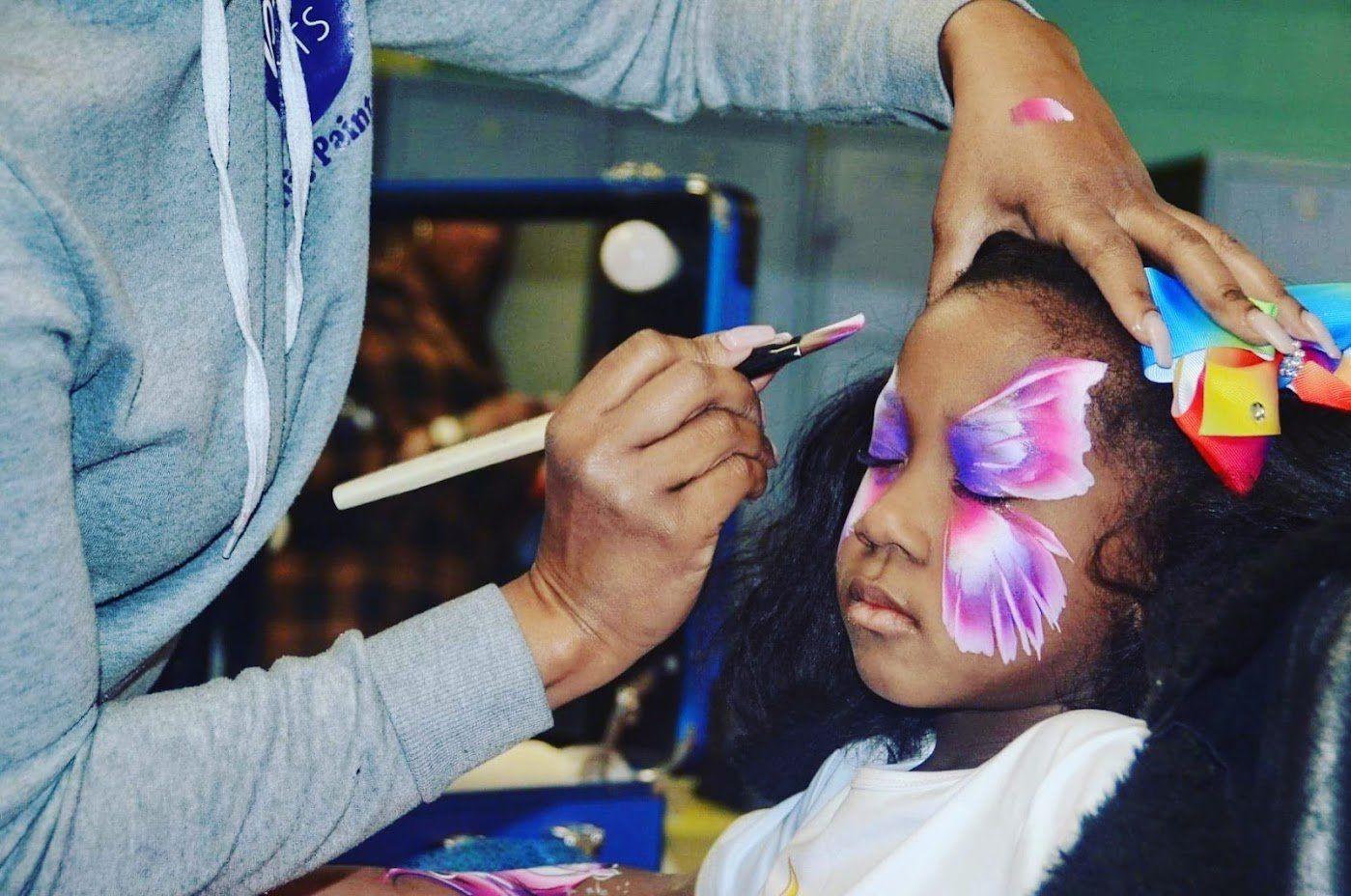 Face painter applying pink and blue butterfly makeup to a child at a table