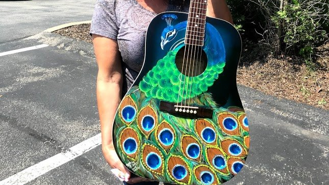 Colorful peacock-themed acoustic guitar with blue, green, and gold feather design, held outdoors on a road