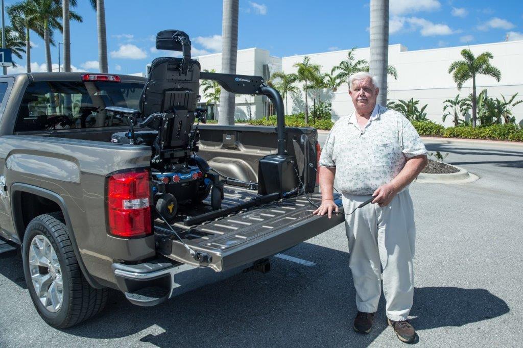 Man standing beside a pickup truck with a wheelchair lift in the bed, outdoors in a parking lot