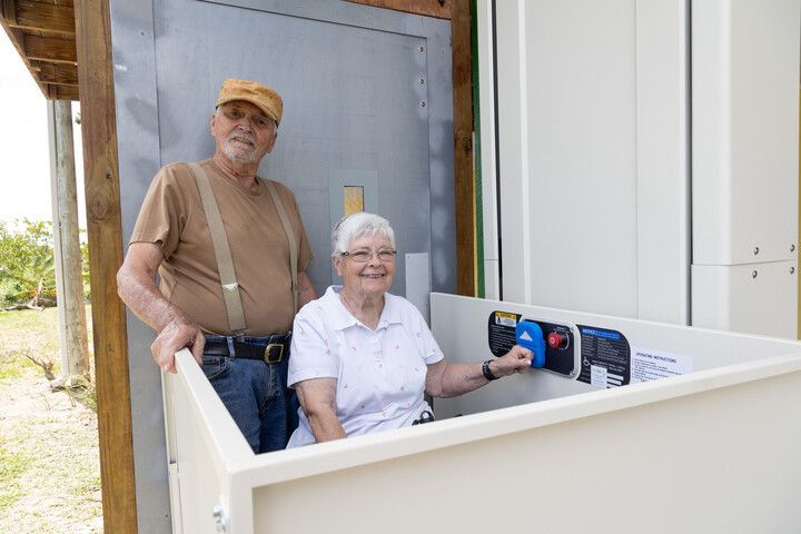 Two people posing in a white accessible lift platform by a doorway, one seated and one standing nearby