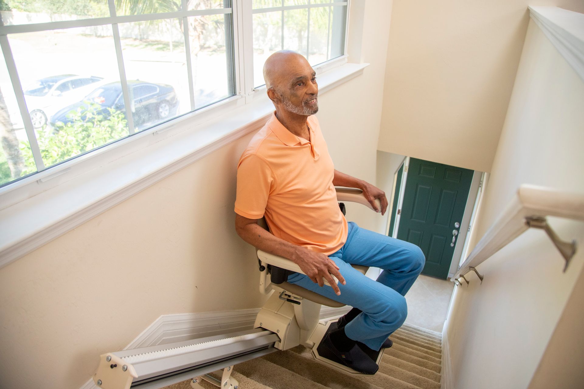 Man in orange shirt riding a stairlift down a narrow staircase by a window