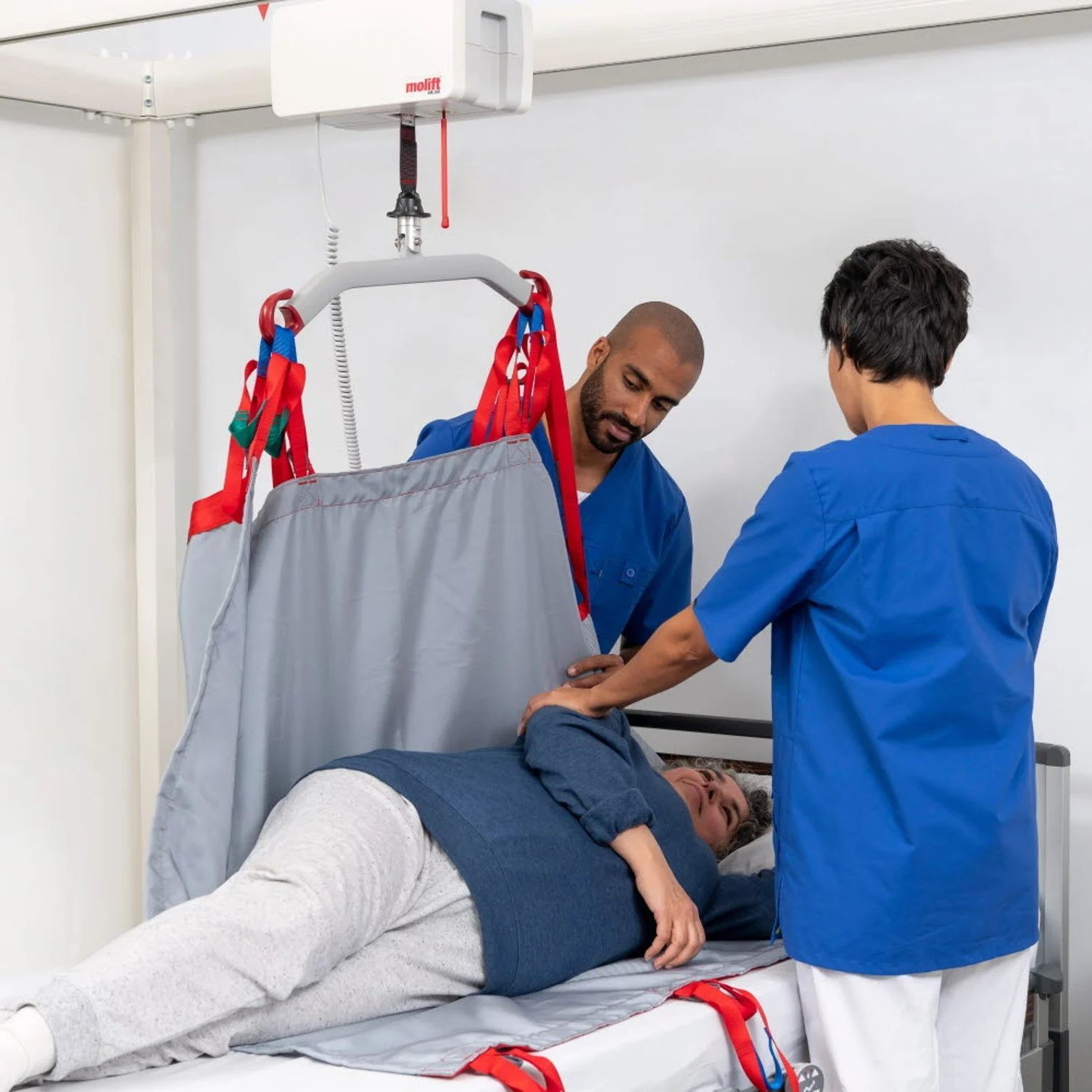 Caregivers transfer a patient on a hospital bed using a ceiling lift sling.