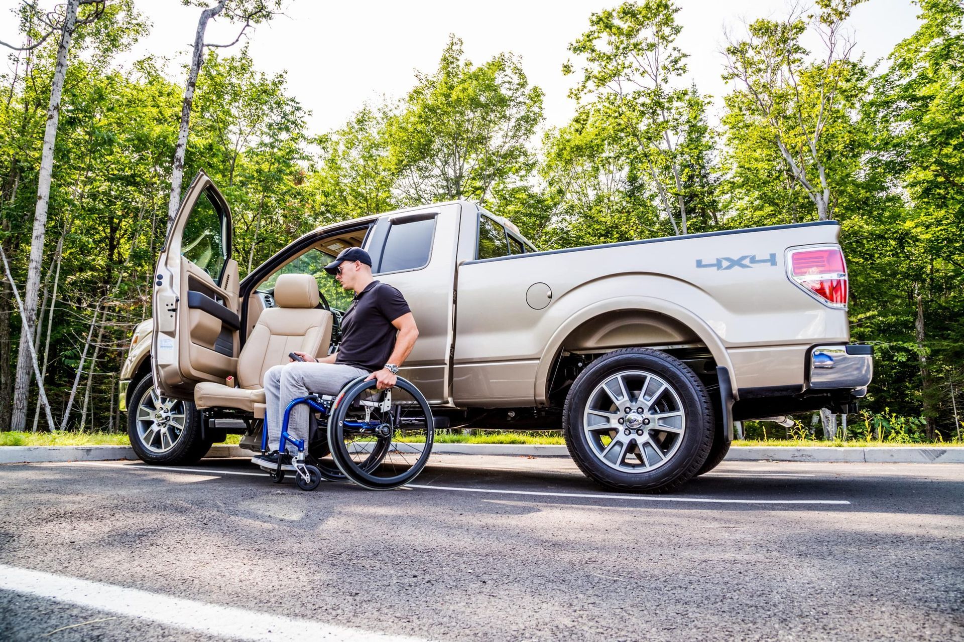Man in wheelchair loading into tan pickup truck on a roadside, with a woman in the driver’s seat
