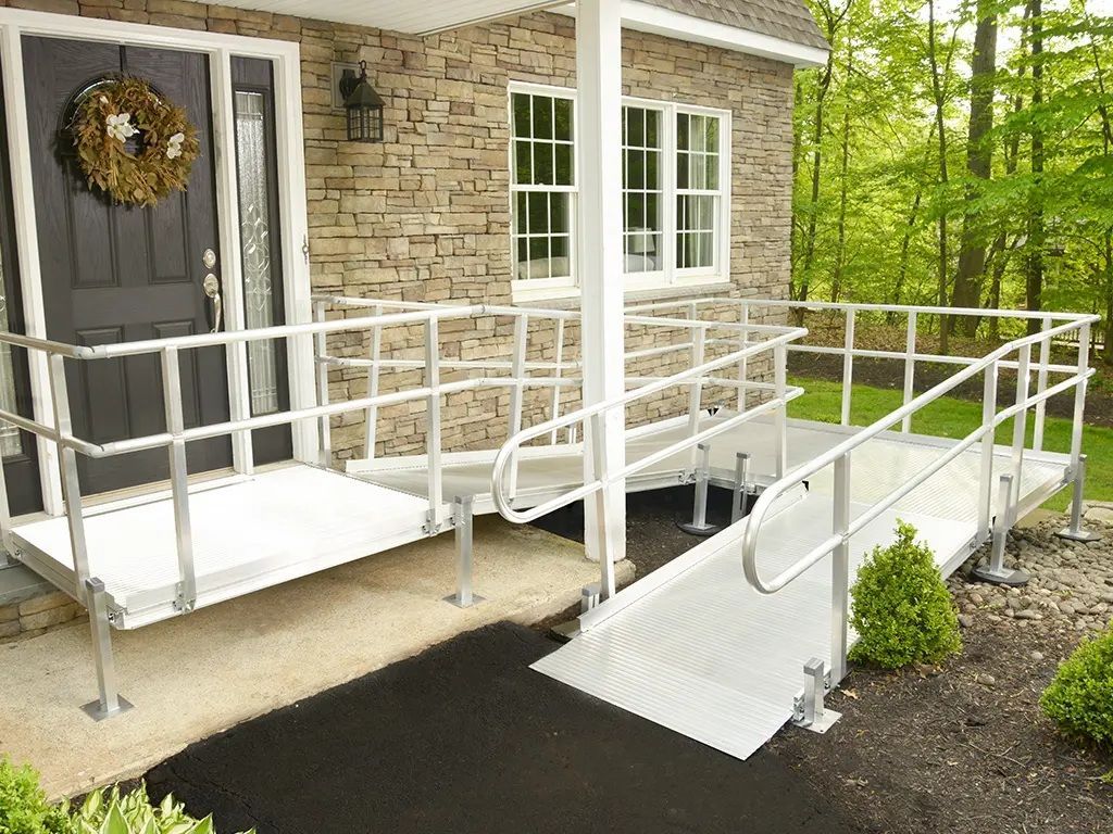 House entrance with wheelchair ramp, white railings, and black walkway beside stone siding and wooded yard