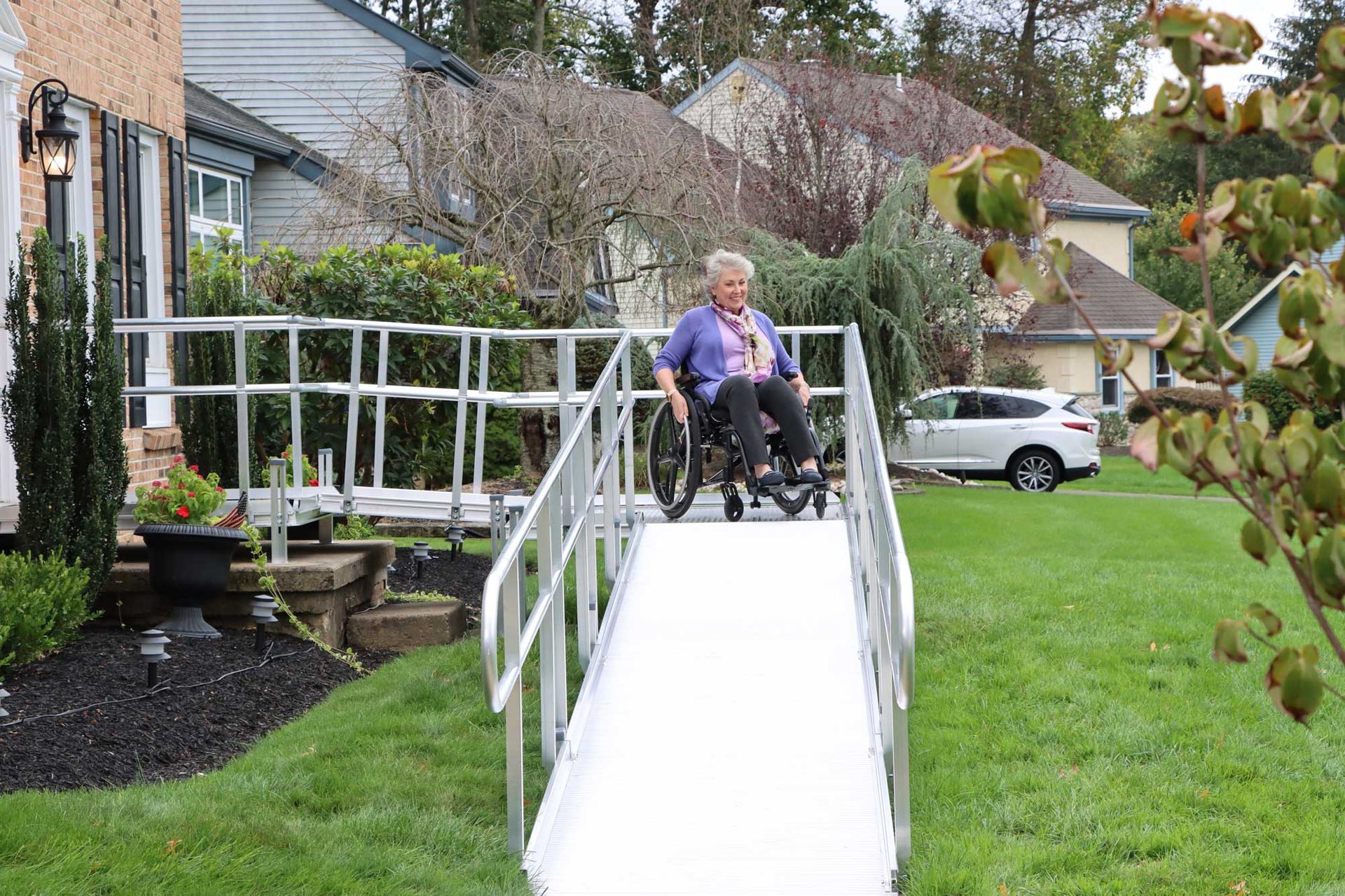 Person in a wheelchair on a white ramp beside a house, with a parked car and grassy yard in the background