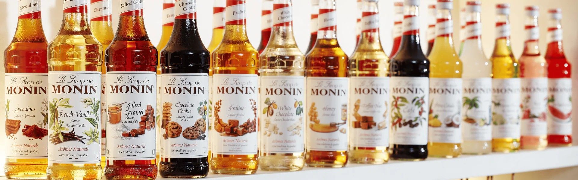 Une rangée de bouteilles de sirop Monin colorées, aux saveurs fruitées assorties, est présentée sur une étagère blanche.