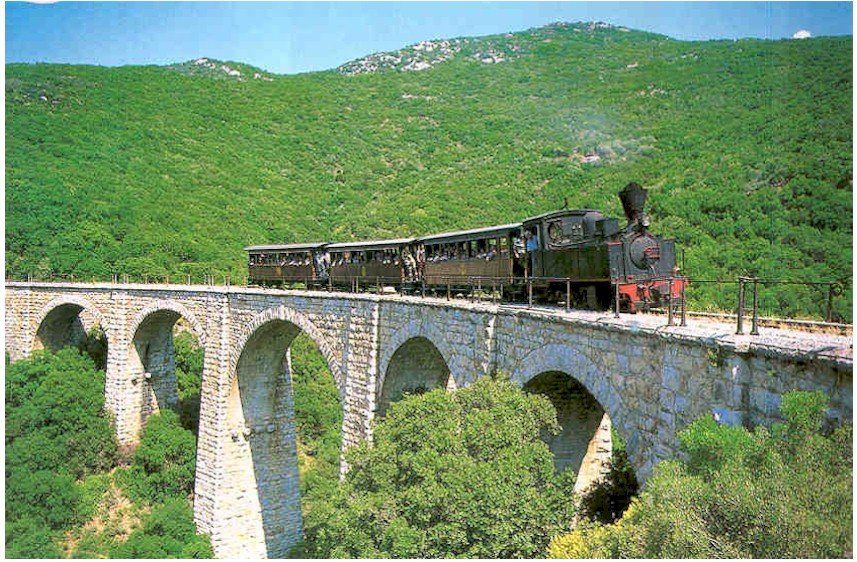 Steam train on a stone bridge in a green landscape, trees, and hills visible.