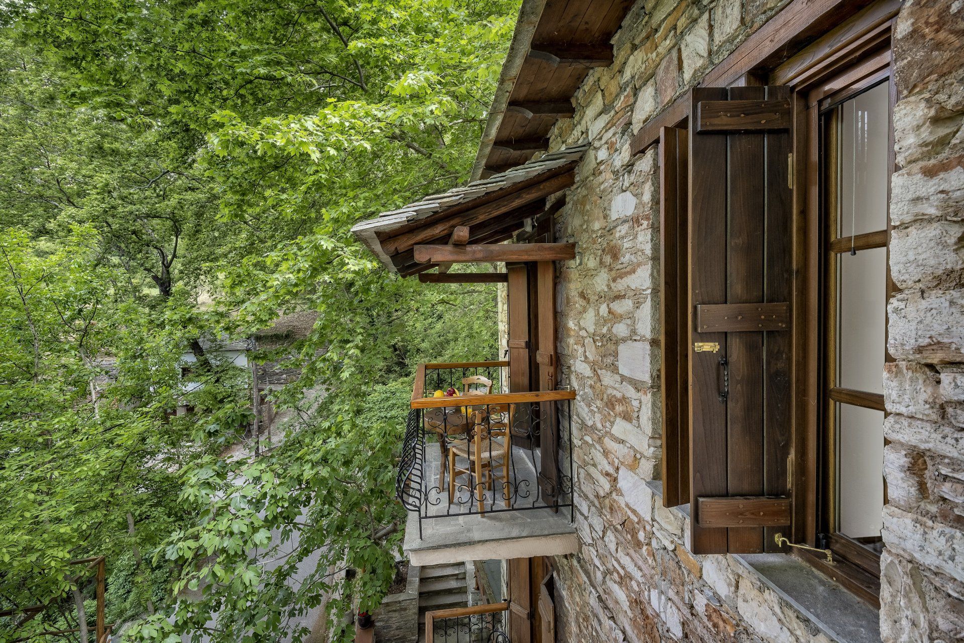 Stone building with wooden shutters and balcony, surrounded by lush green foliage.
