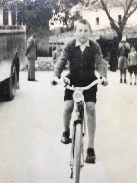 Boy riding a bicycle, looking forward. Wearing dark shorts and a sweater. Outdoors with people and a bus.
