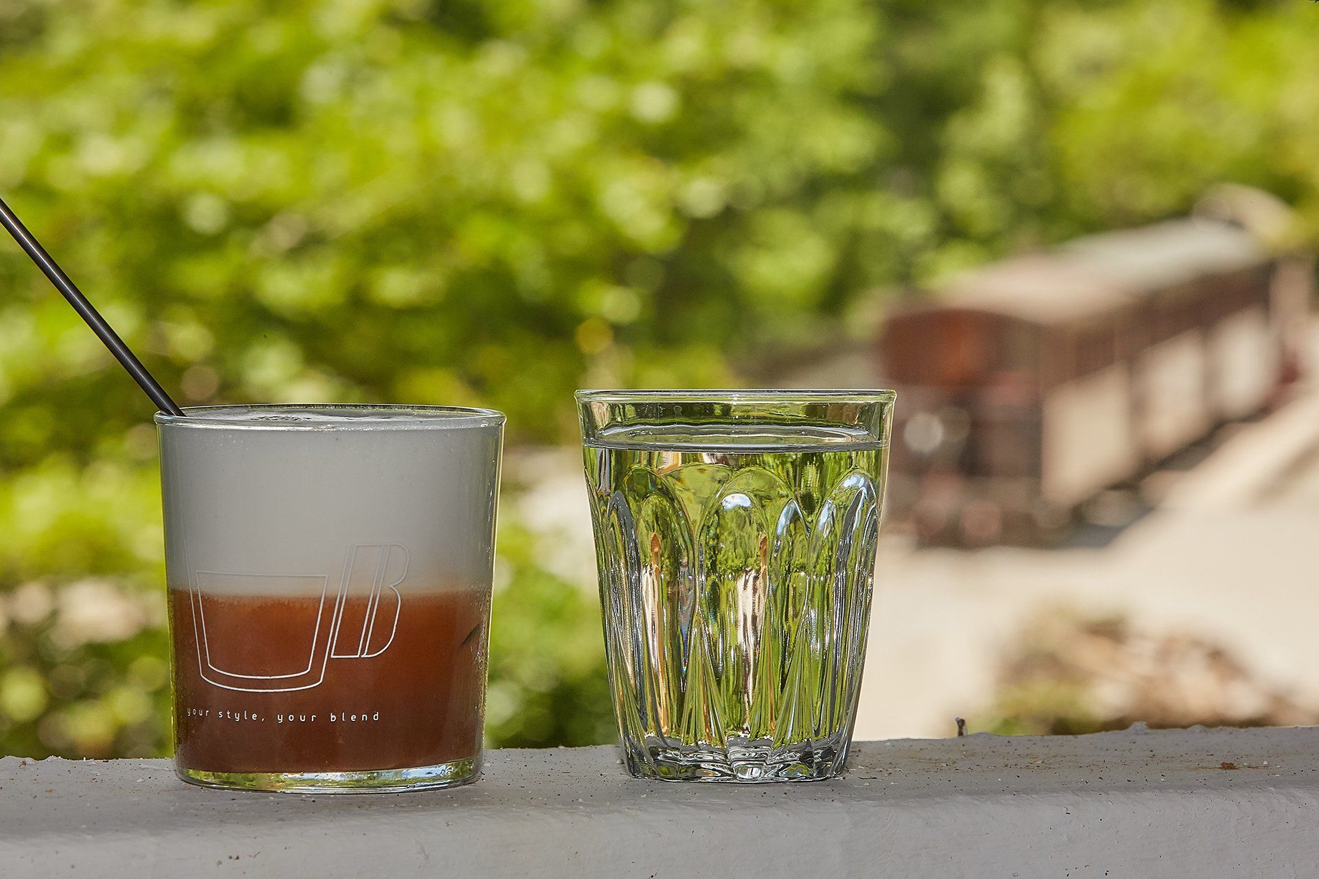 Two glasses on a white surface; one with a brown drink and foam, the other with clear liquid. Blurry green background.