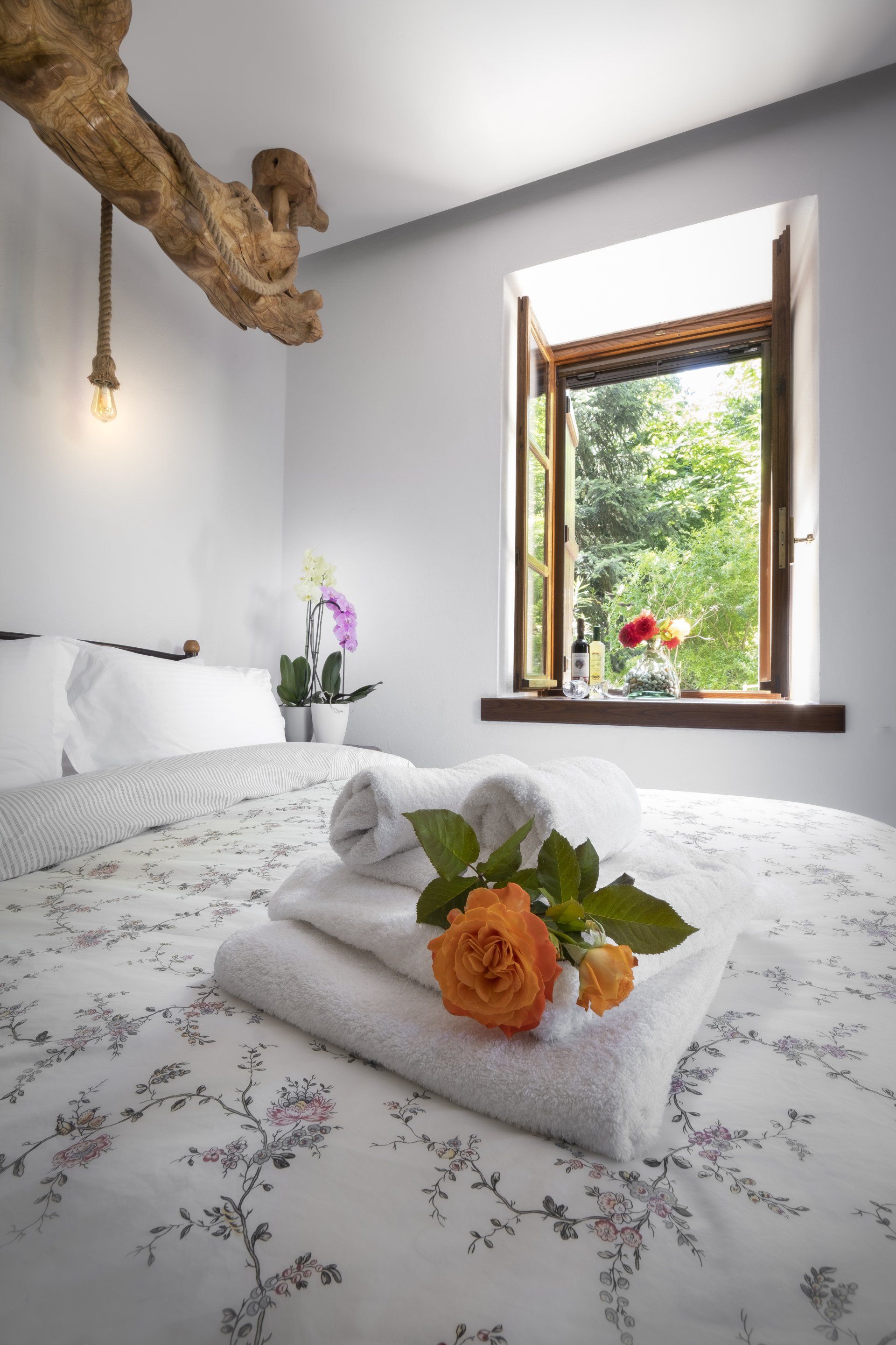 Bedroom with a bed covered in floral linens, white towels, an orange rose, and an open window with greenery outside.