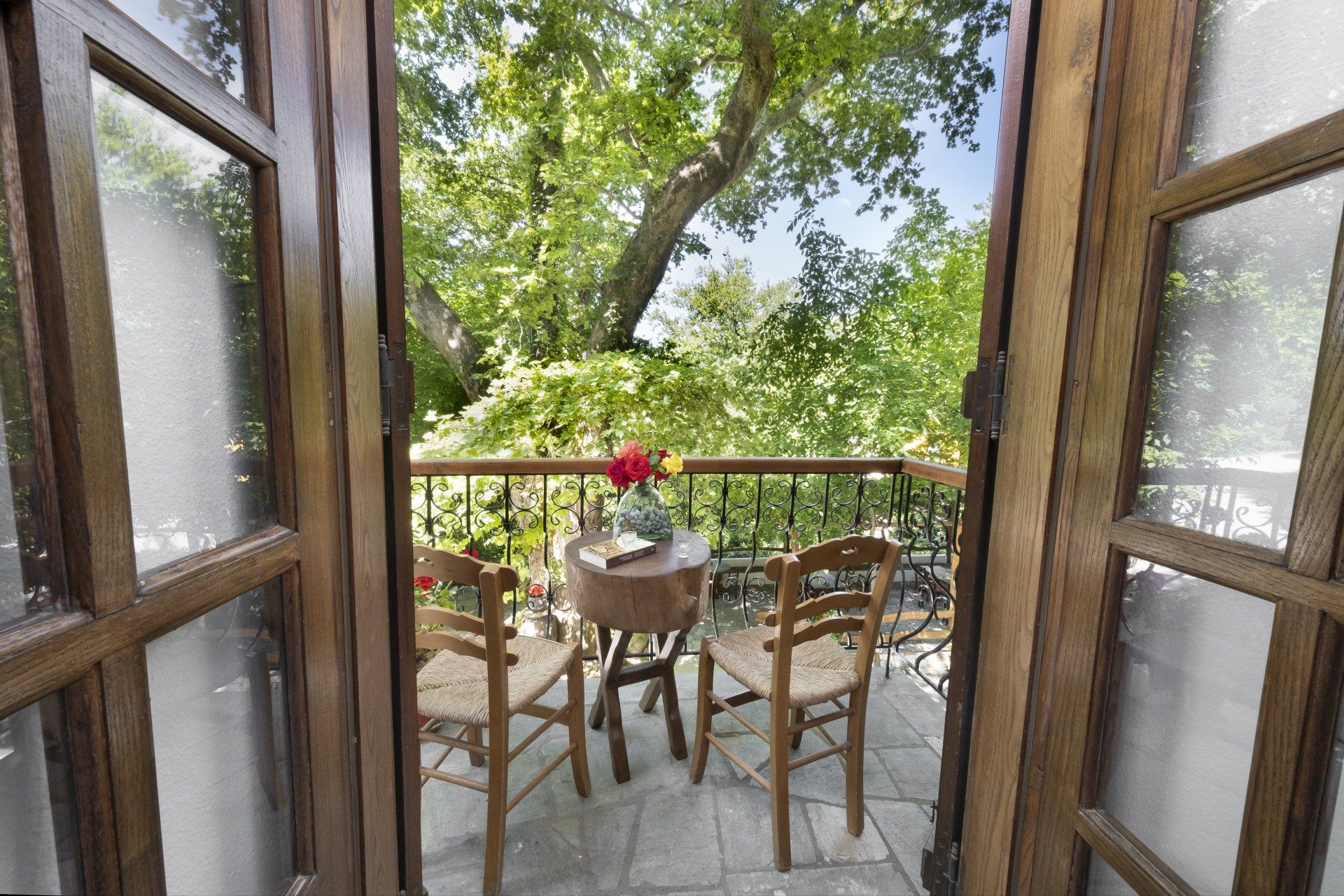 Wooden balcony with two chairs, small table, and open doors with a view of greenery.