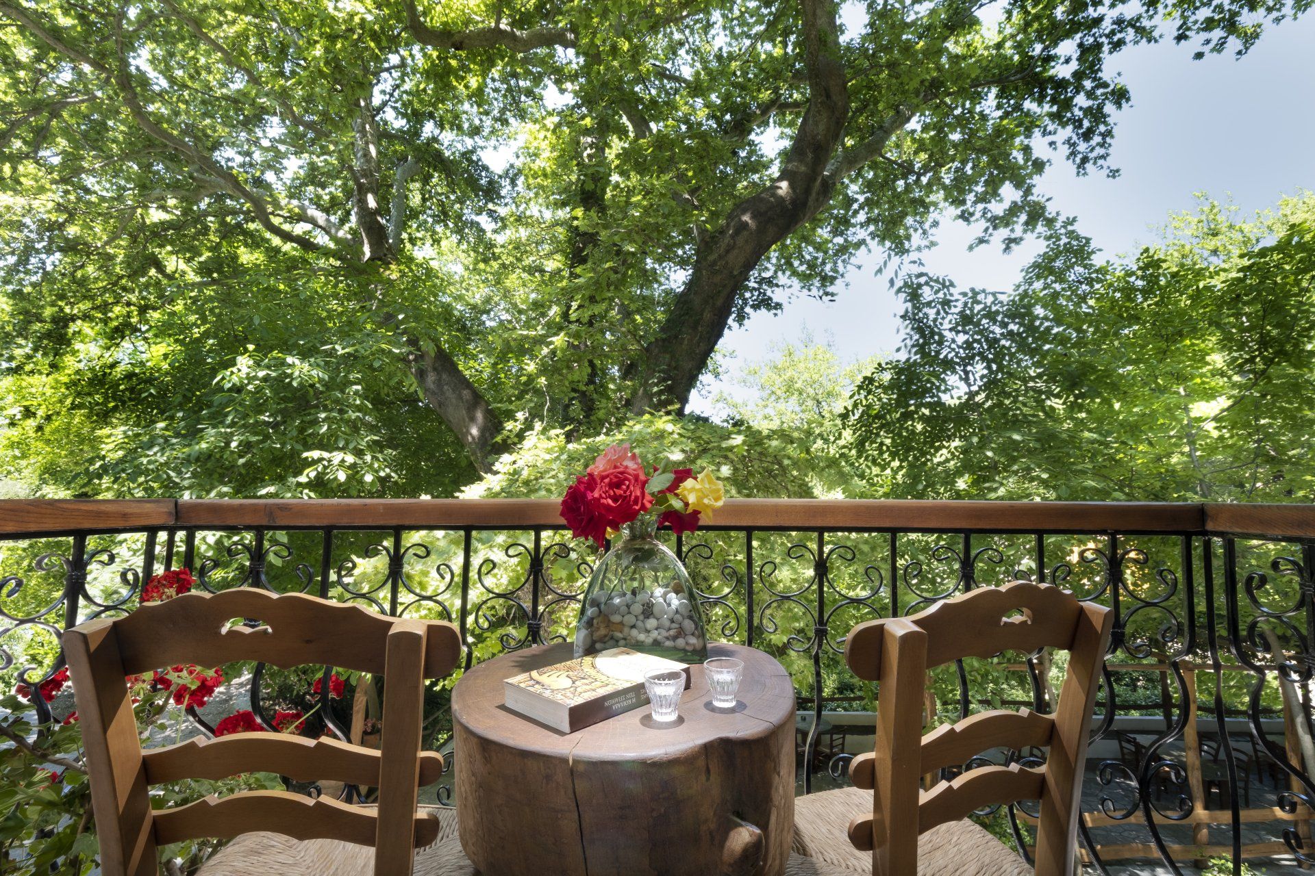 Balcony with wooden chairs, small tree-stump table, vase of flowers, book, and expansive green tree backdrop.