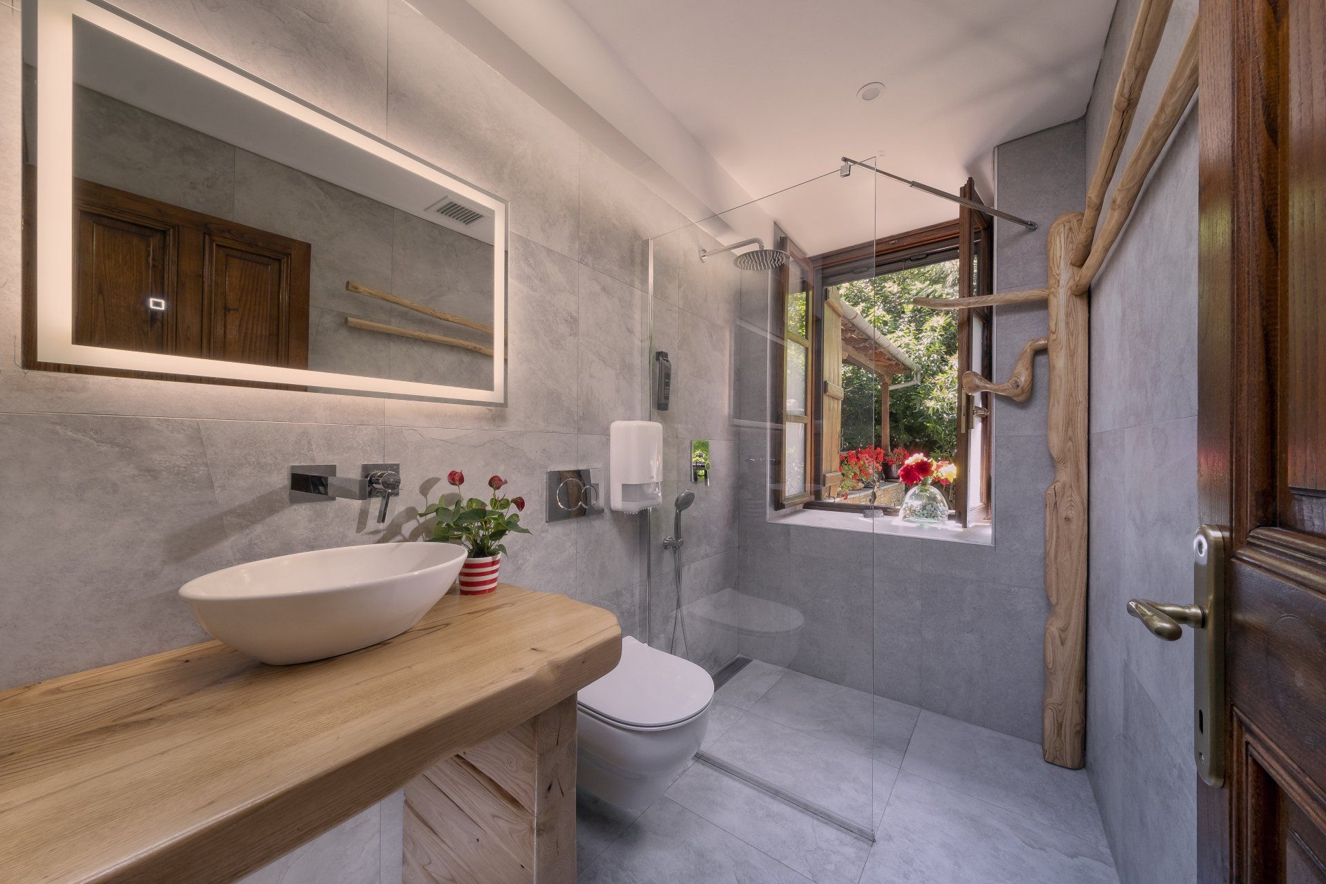 Bathroom with a wooden vanity, gray walls, white sink, toilet, and a window with plants.
