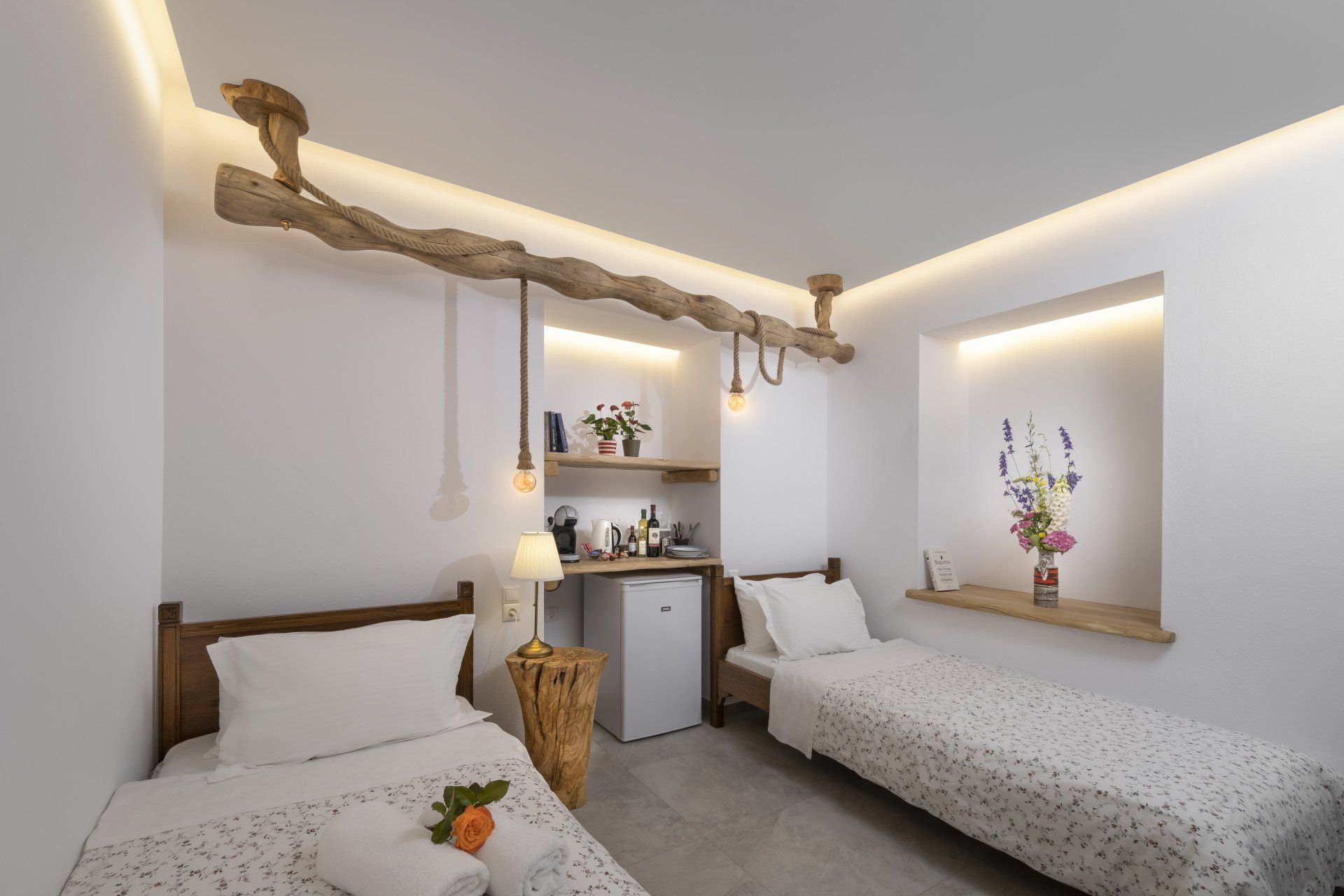 Two twin beds in a white room with rustic wood accents, a small kitchenette, and floral arrangements.