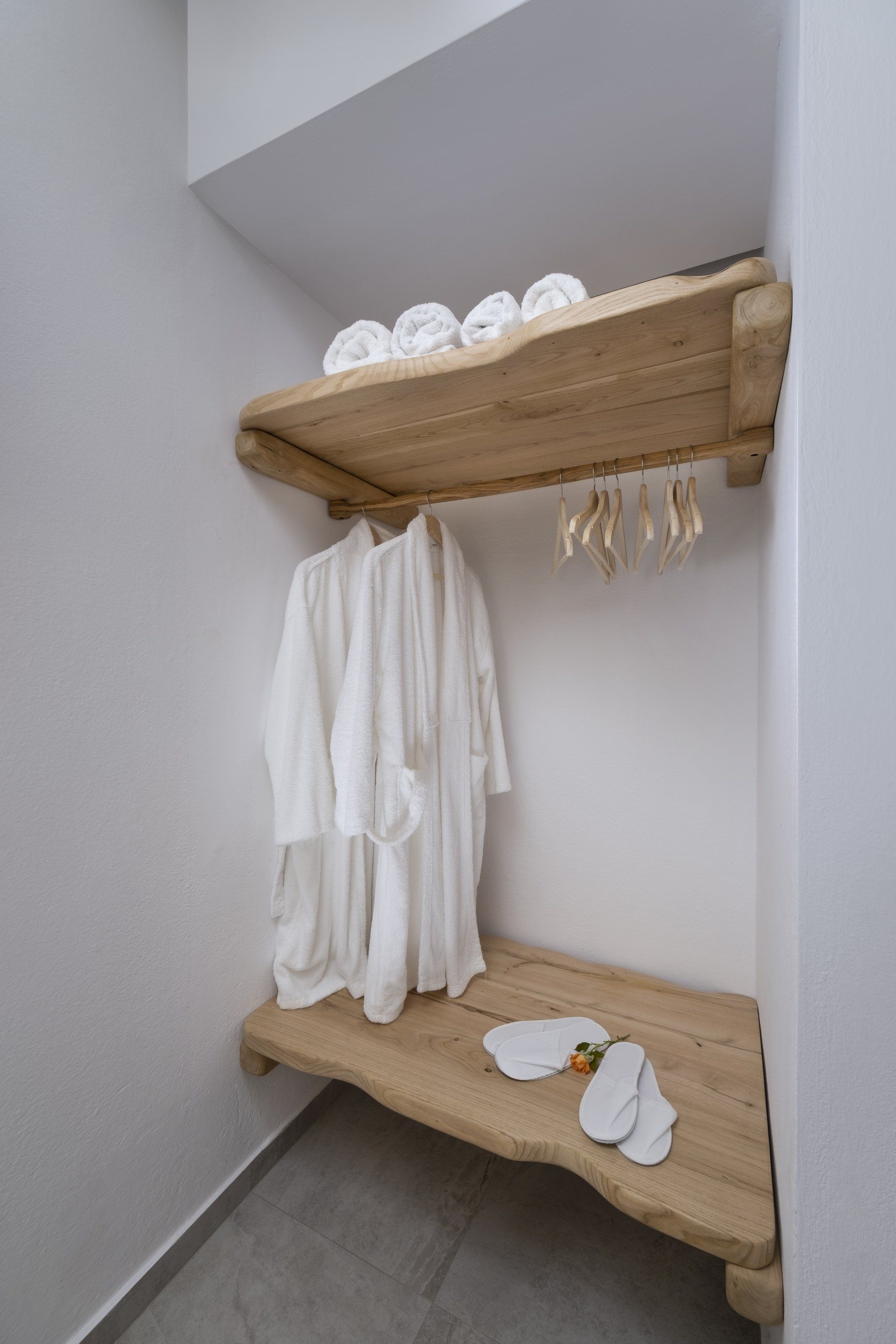 Built-in closet with rough-hewn shelves, holding robes, towels, slippers, and hangers against a white wall.