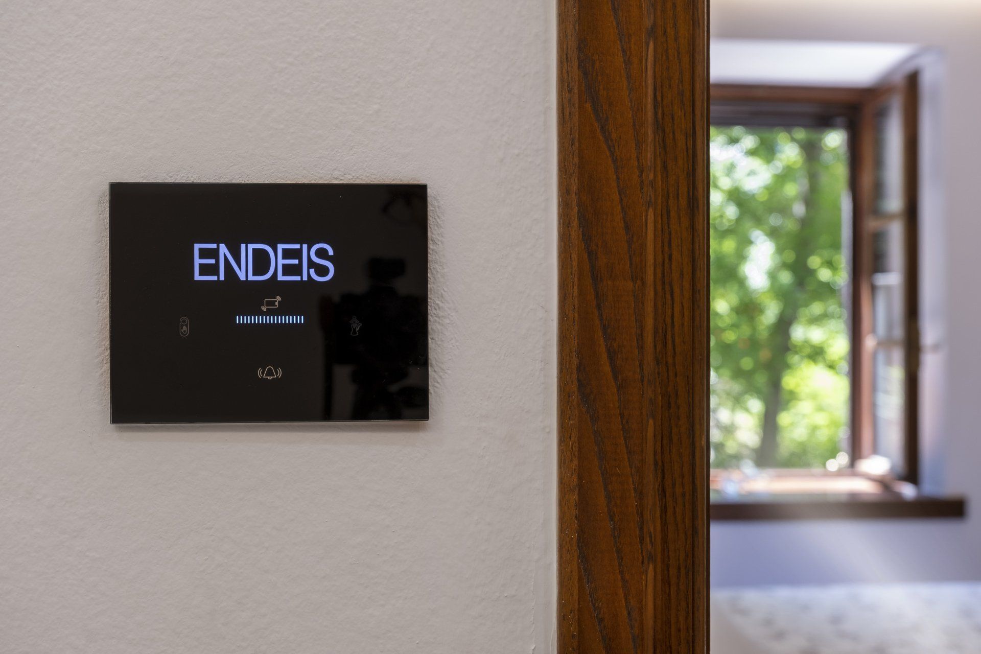 Black smart home control panel on white wall, wooden door frame, open window with foliage.