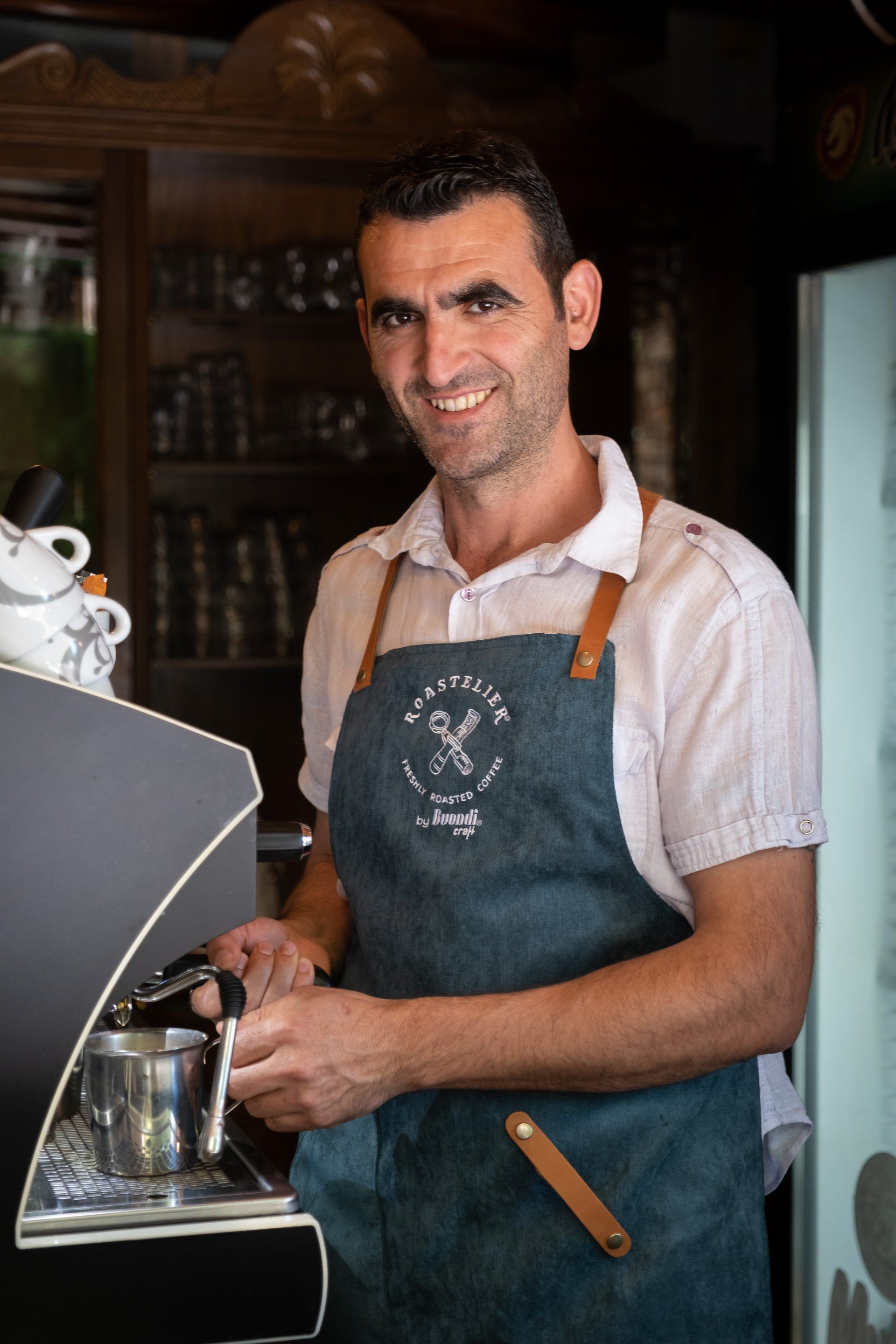 Man in apron prepares coffee at a machine, smiling. Indoor setting.