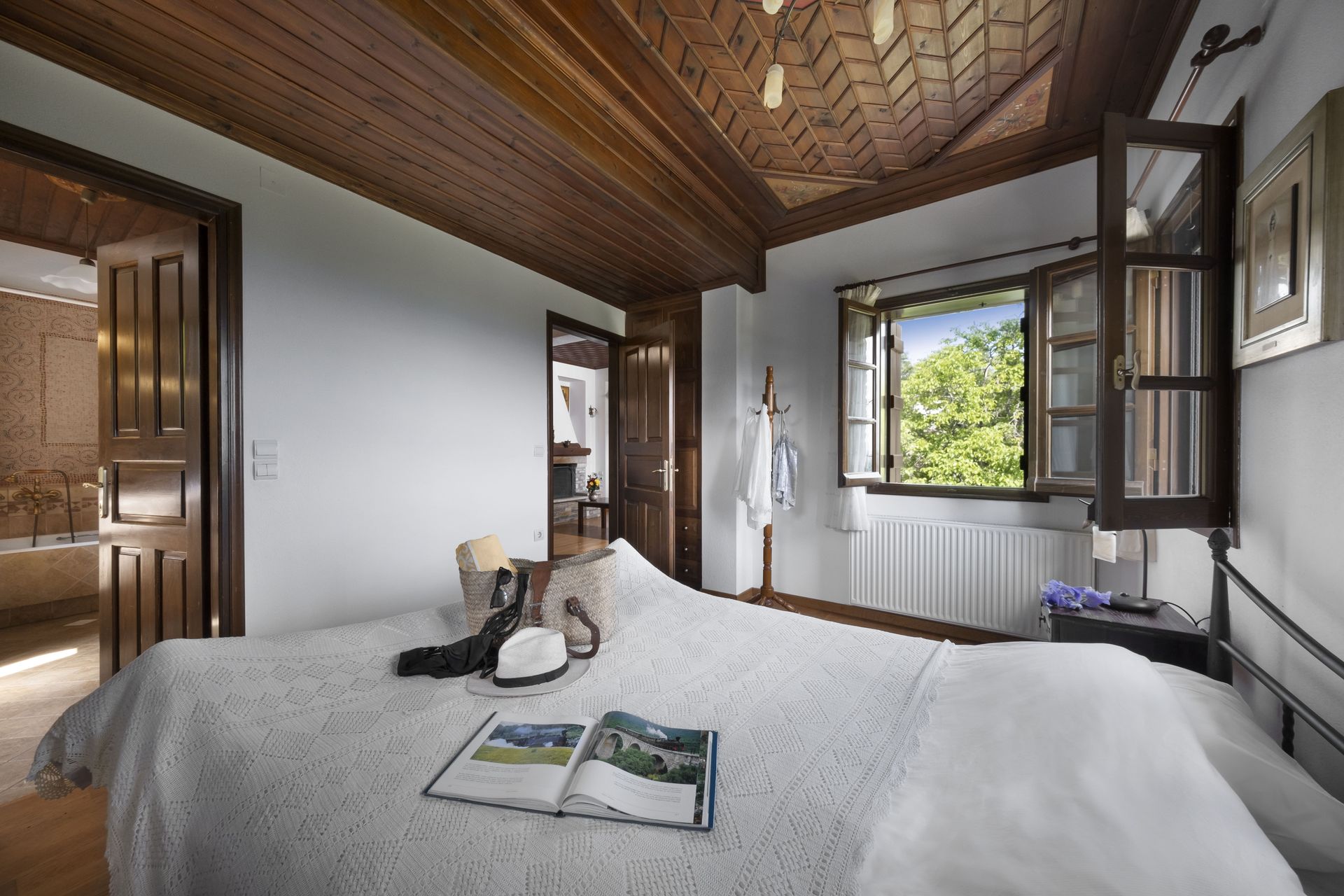 Bedroom with wooden ceiling, open window overlooking greenery, bed with white cover, open book.