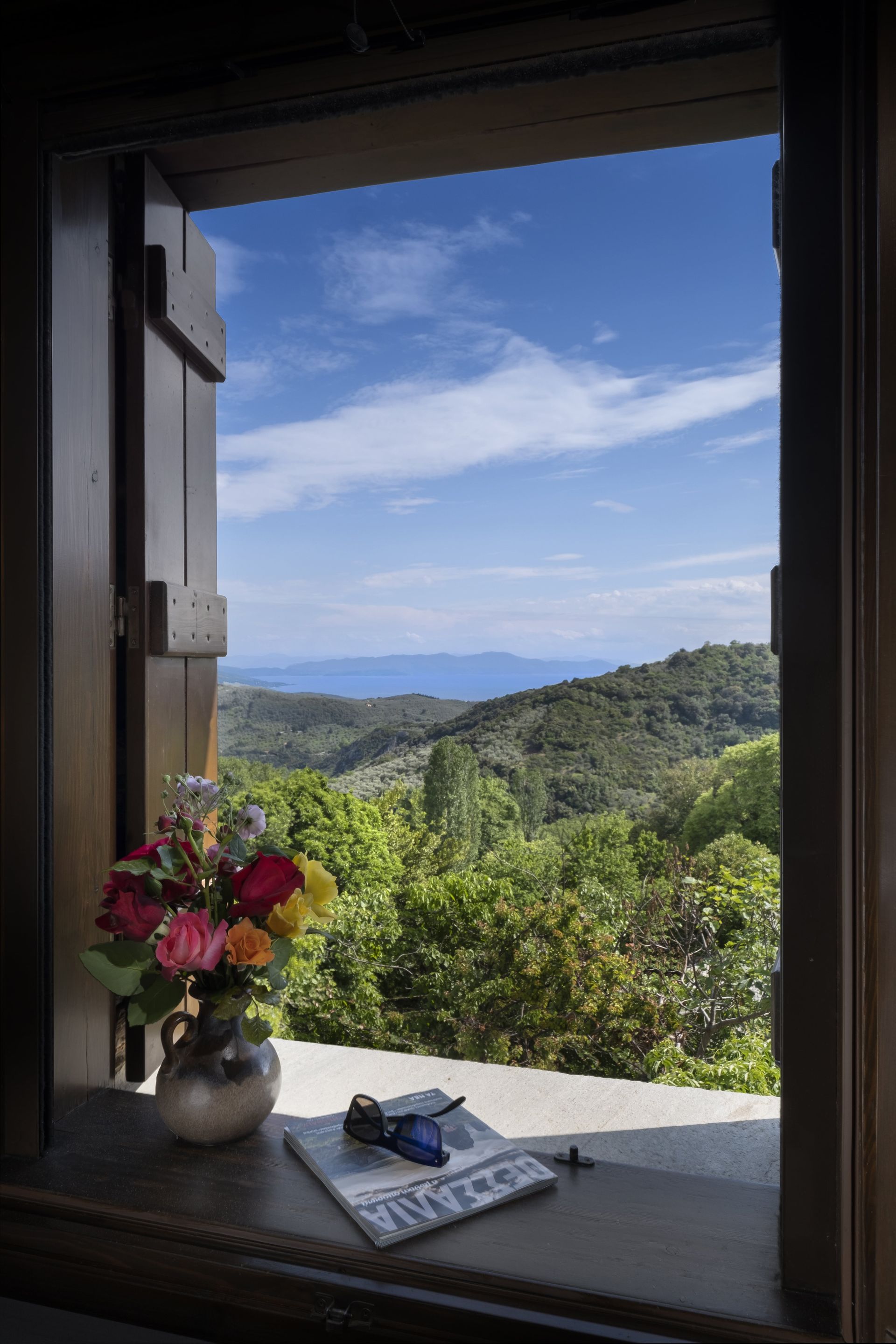 Window view of green hills, blue sea, and sky. Flowers, glasses, and paper on the sill.