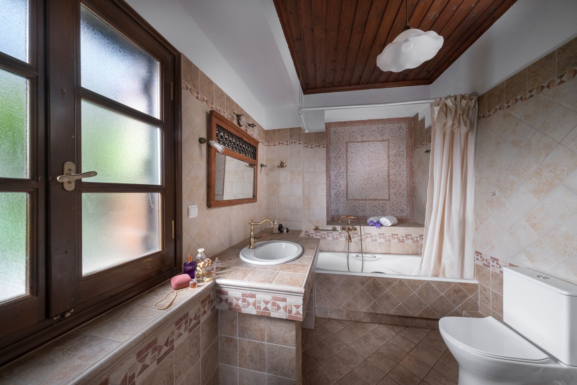 Bathroom with window, sink, toilet, and bathtub. Beige tiled walls, wooden ceiling, and curtain.