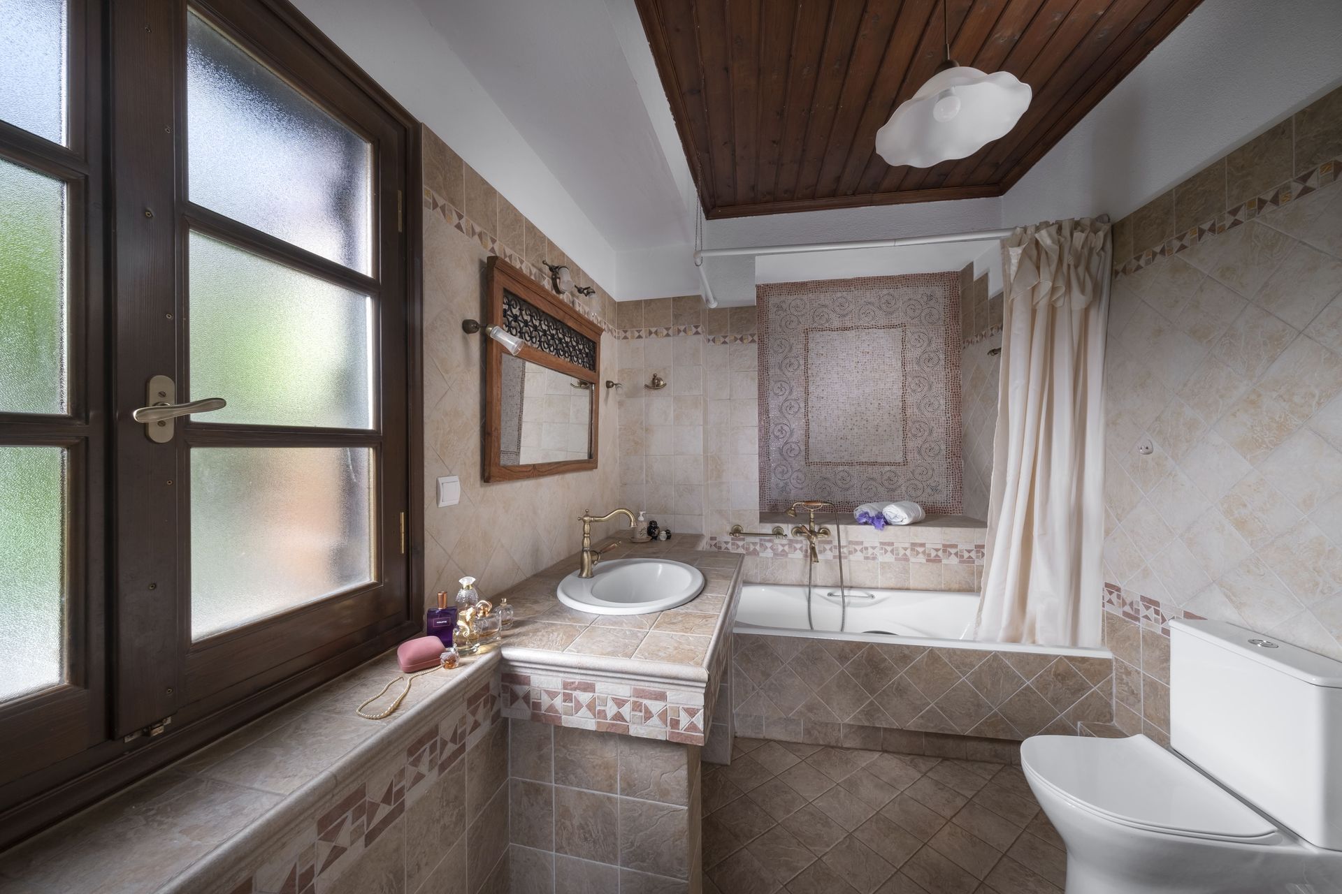 Bathroom with window, sink, toilet, and bathtub. Beige tiled walls, wooden ceiling.