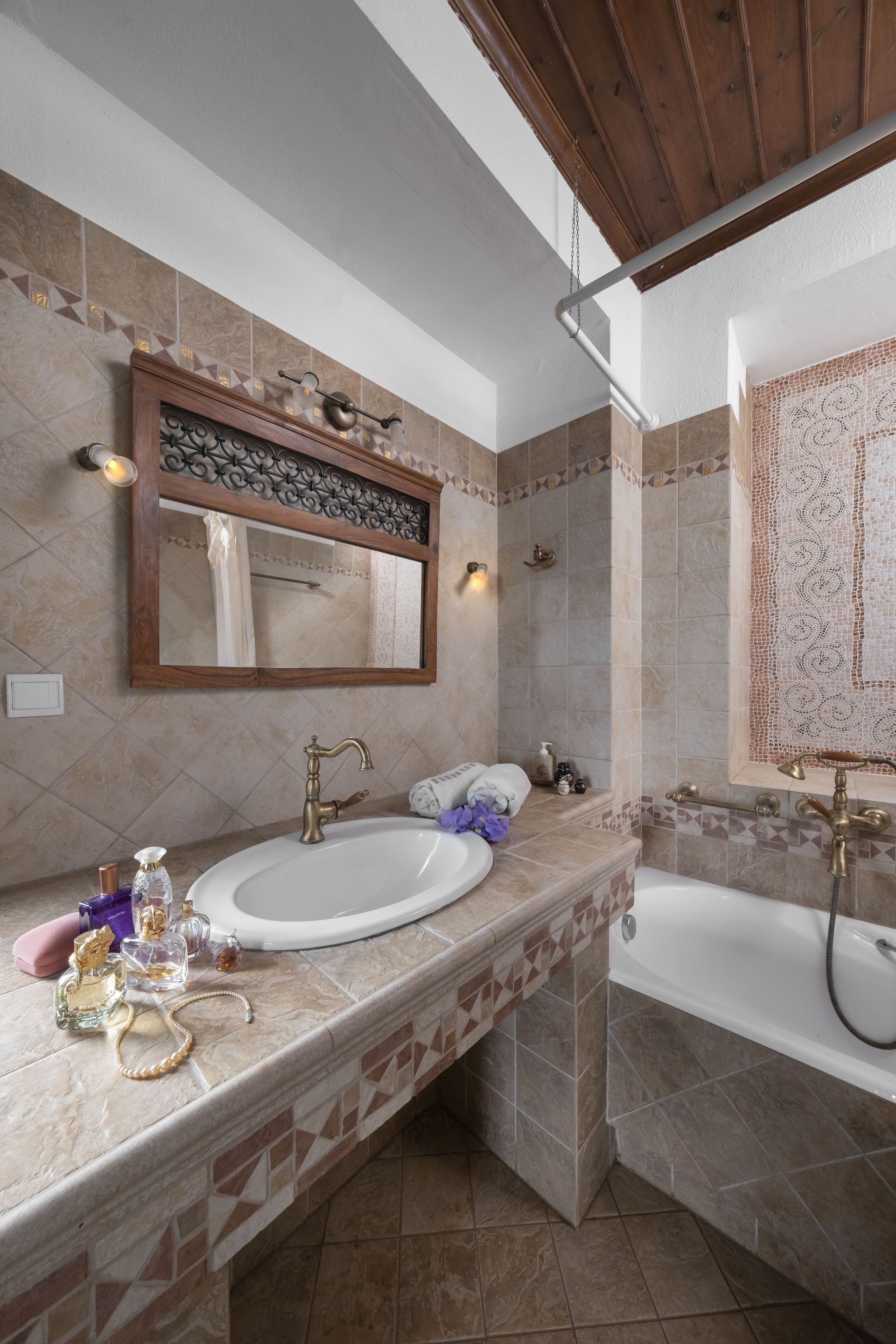 Bathroom with a sink, mirror, and bathtub; beige tiles and a wooden ceiling give a rustic vibe.