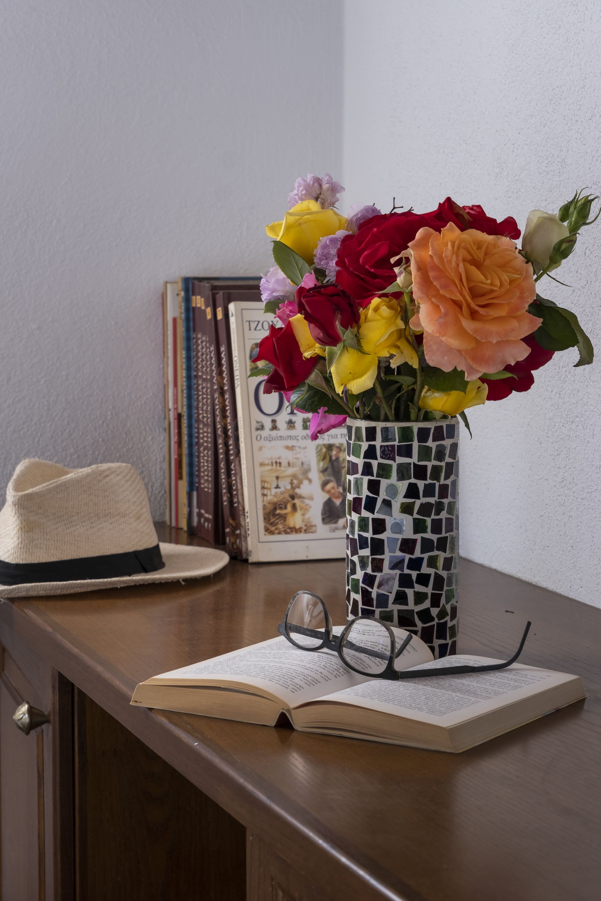 A bouquet of colorful roses in a mosaic vase, book with glasses, hat, and books on a wooden surface.
