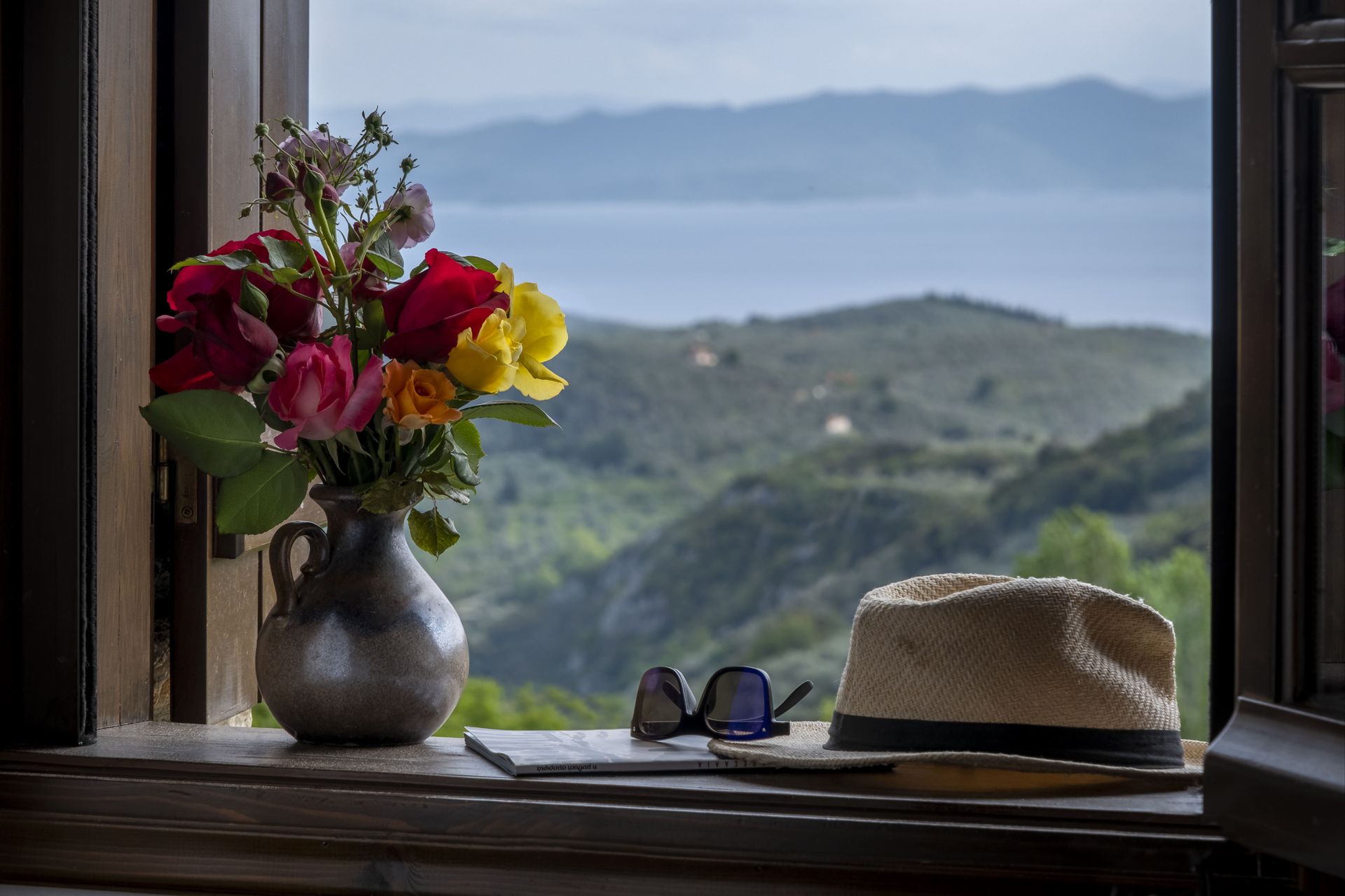 Flowers in a vase, hat, and sunglasses on a windowsill overlooking a green mountain range and ocean.