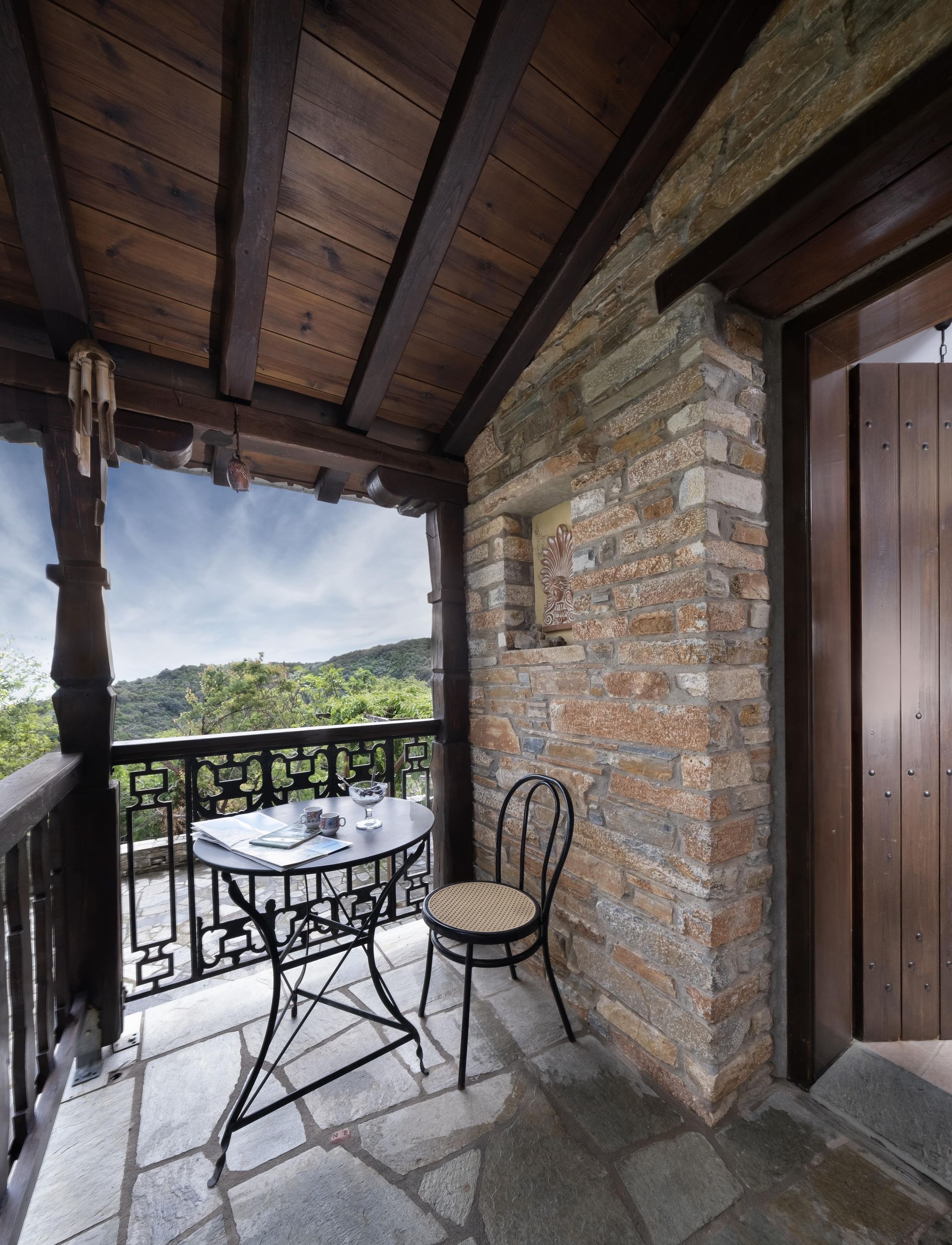 Balcony with table, chair, and view of green hills. Stone wall and wooden beams.