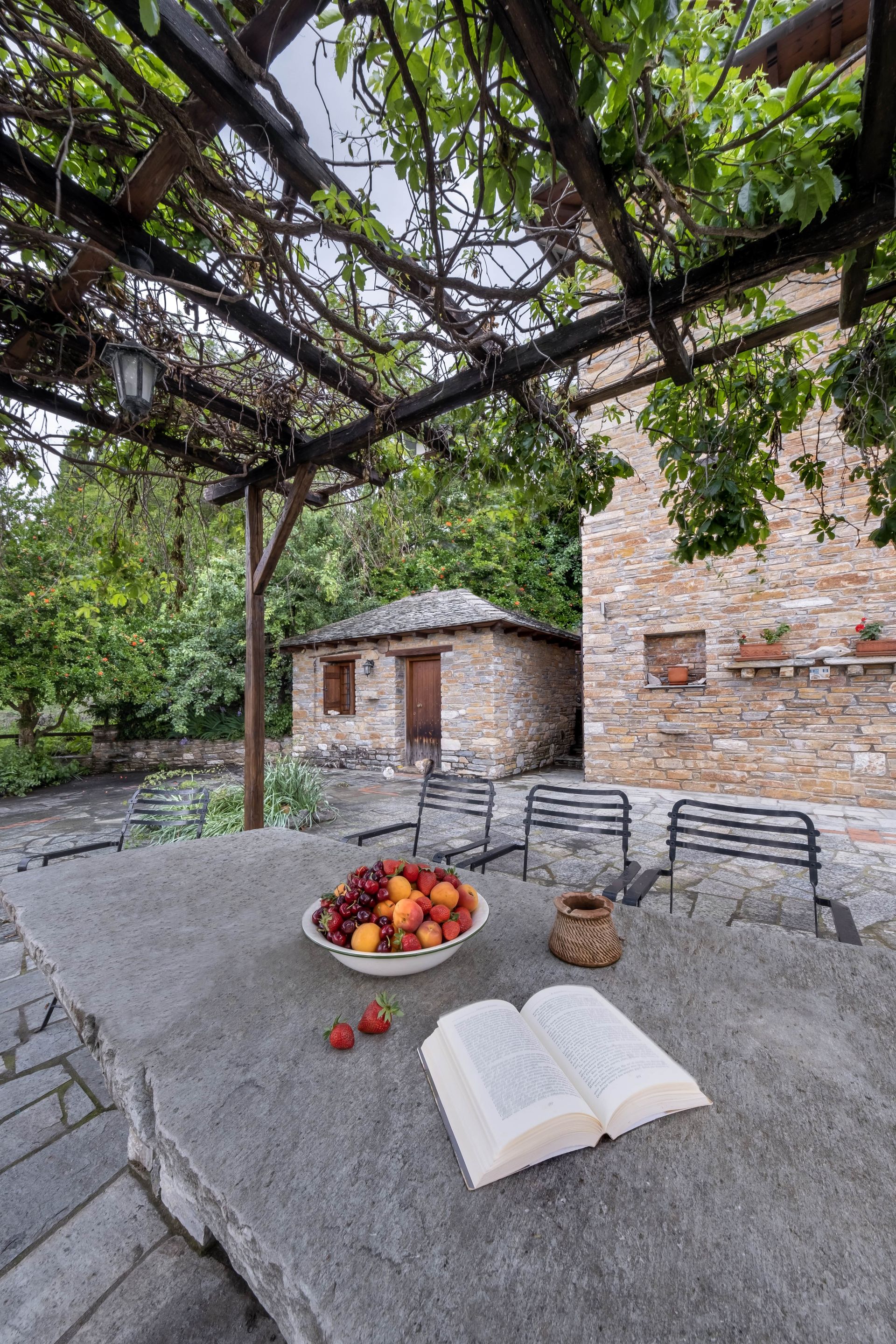 Stone patio with fruit bowl, book, and chairs under a vine-covered pergola, near stone buildings.