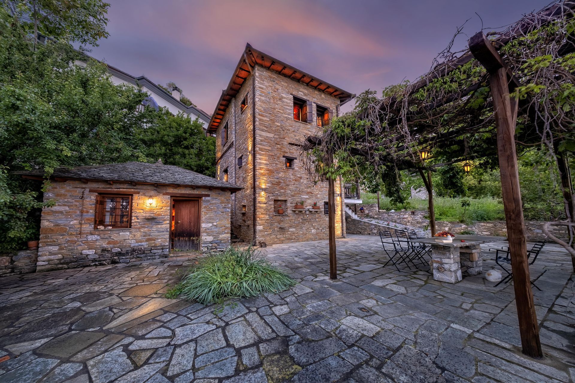 Stone buildings with outdoor seating under a vine-covered pergola at dusk.