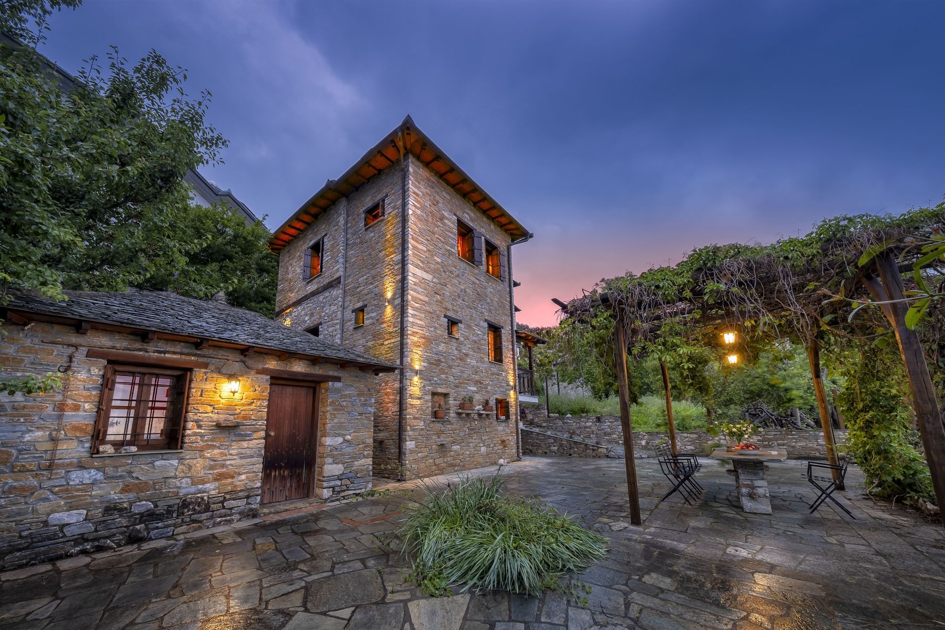Stone building with tower lit at dusk, patio with vine-covered pergola and table, forest background.