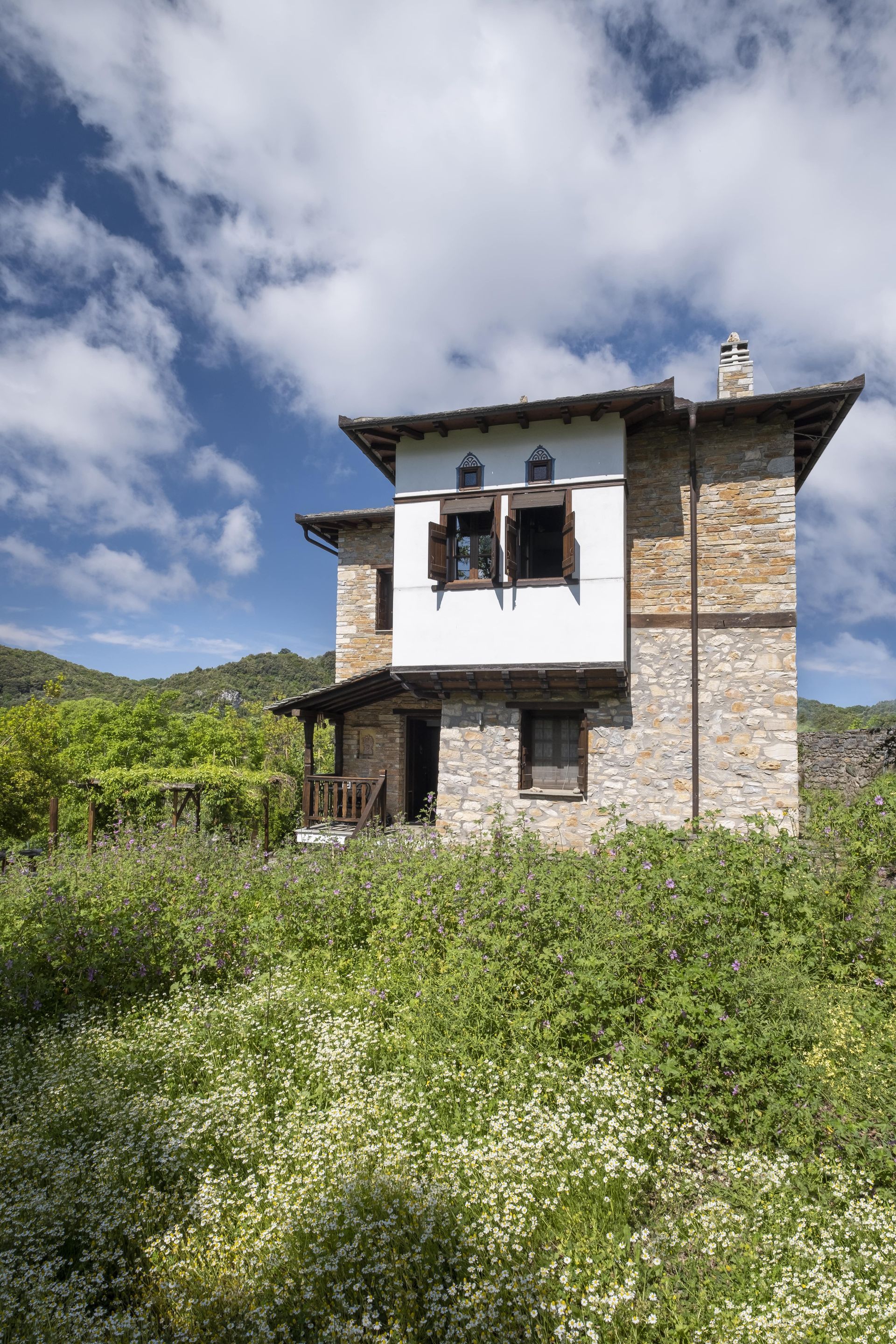 Stone house with white balcony, wooden shutters, set in a field of wildflowers, under a blue sky with clouds.