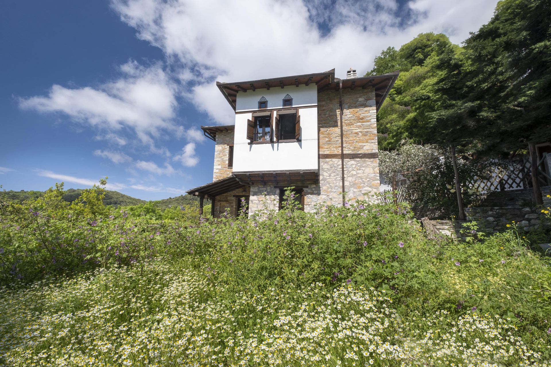 Two-story stone house with white trim, set amidst green vegetation under a blue sky.