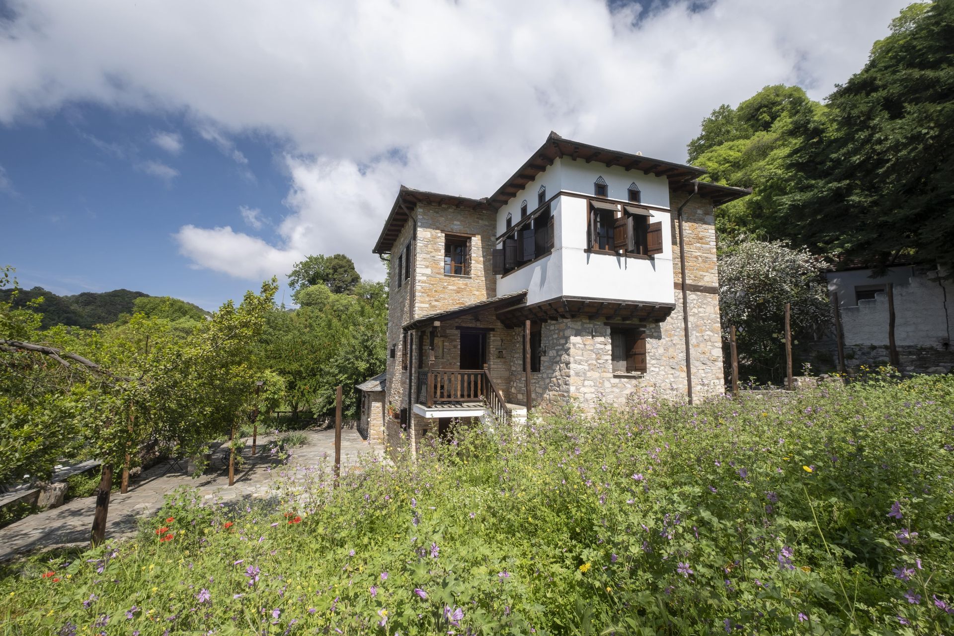 Two-story stone house with white trim and wooden shutters, on a hillside with green foliage and cloudy blue sky.