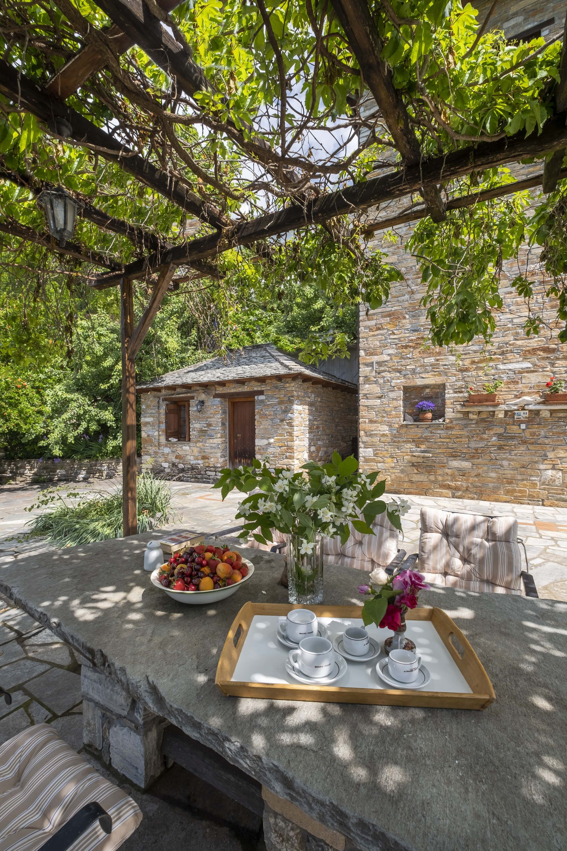 Stone patio with table set for tea, under a vine-covered pergola. Fruit bowl and flower vase sit on the table.