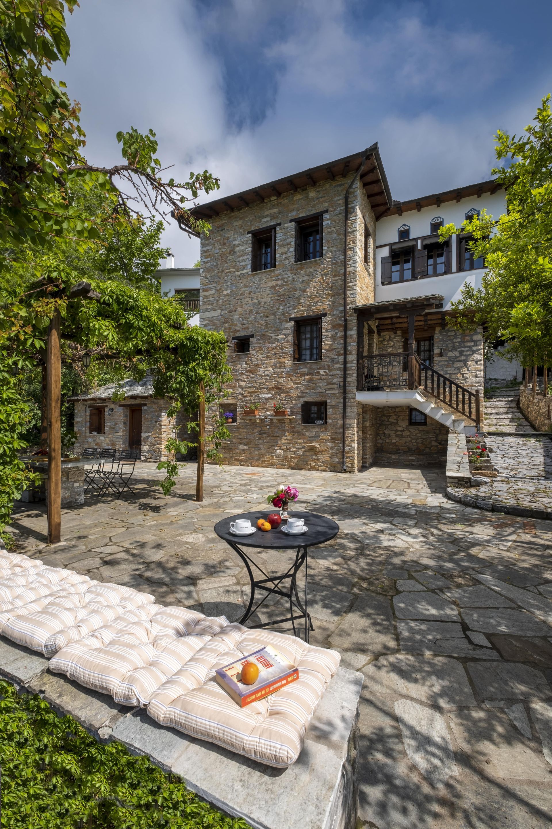 Stone building with outdoor seating area; table with breakfast, bench with cushions, trees overhead.