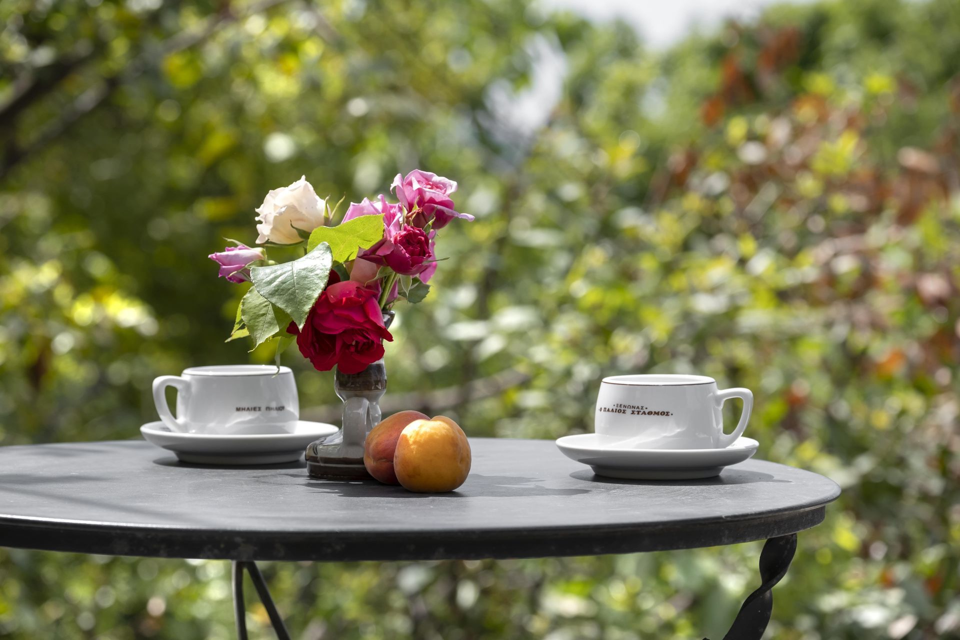 Two coffee cups on a table with a flower bouquet and fruit, set against a blurred green backdrop.