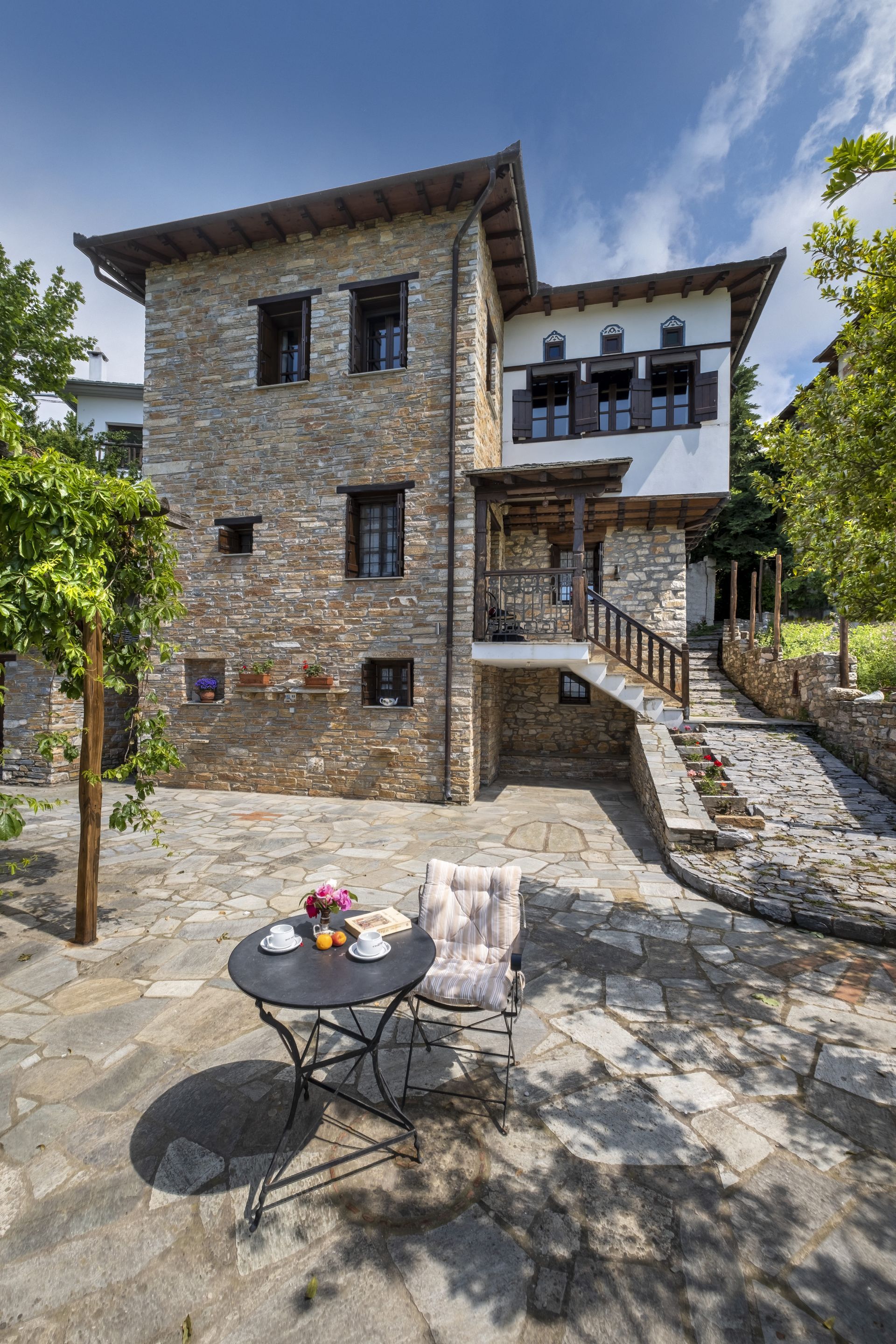 Stone house with balcony, stairs, and patio. Table with fruit and chair in foreground.