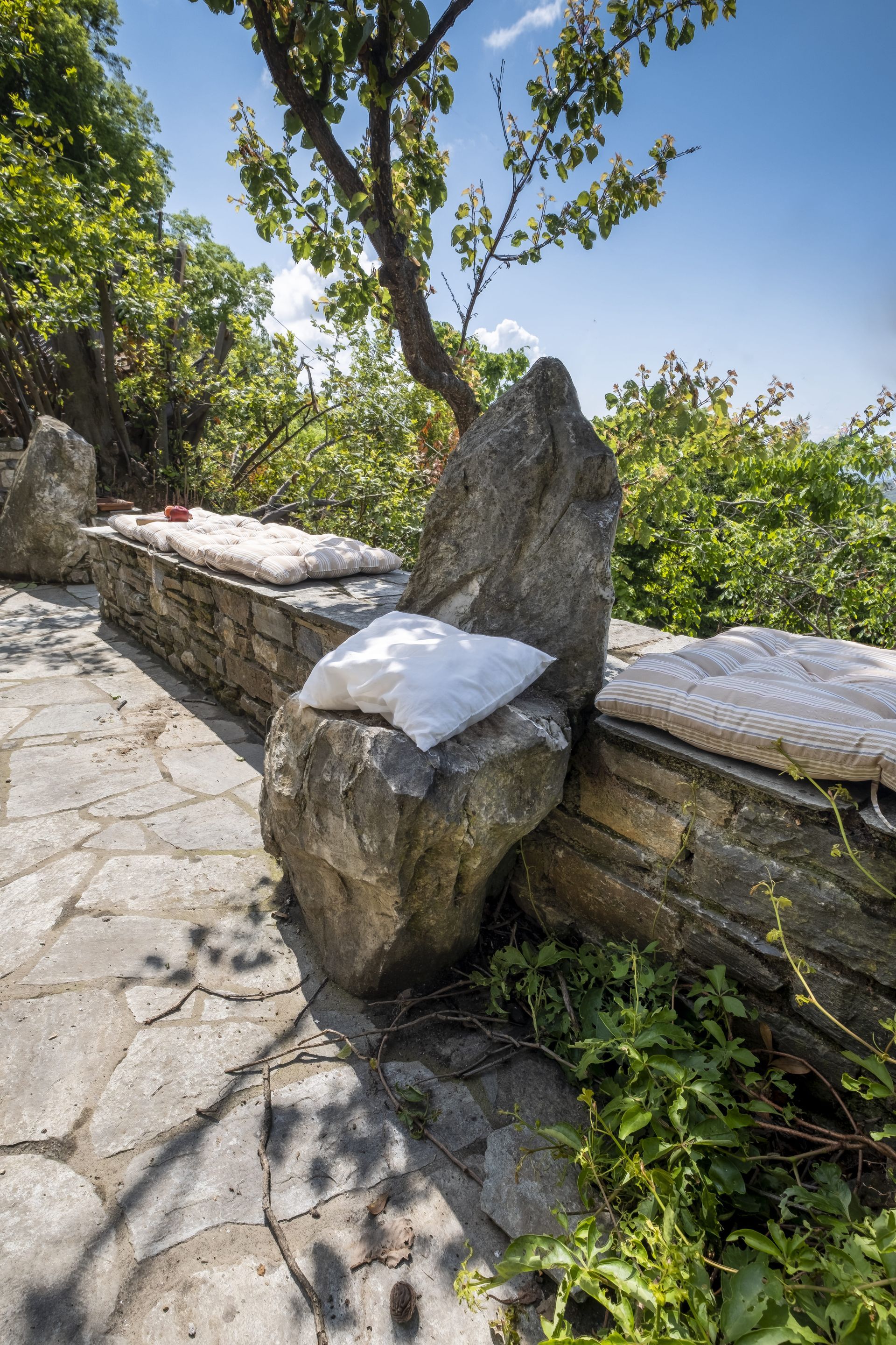 Stone bench with large rock backrest, overlooking greenery on a sunny day.