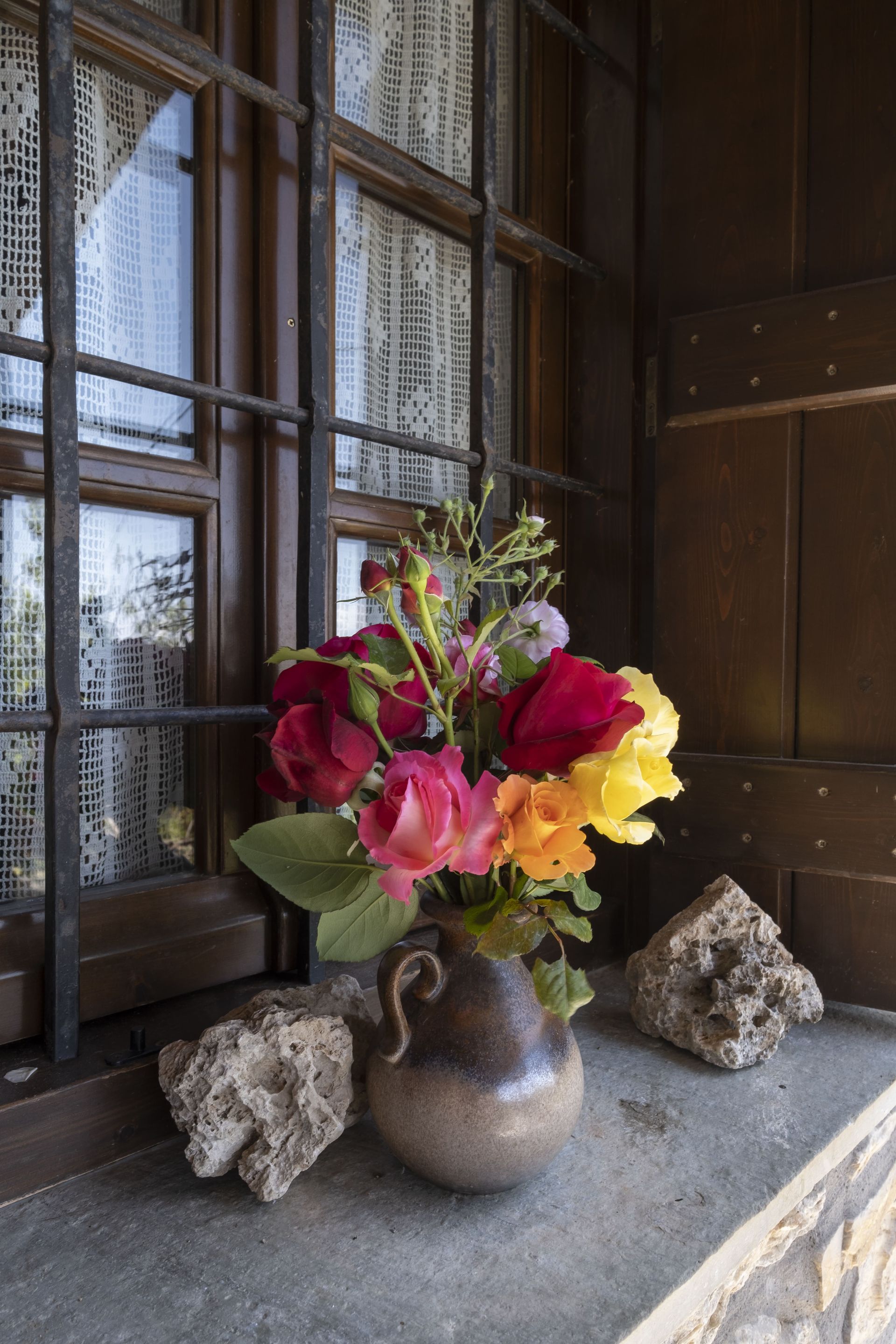 Bouquet of red, pink, and yellow flowers in a ceramic vase on a windowsill, beside two textured rocks, near a window.