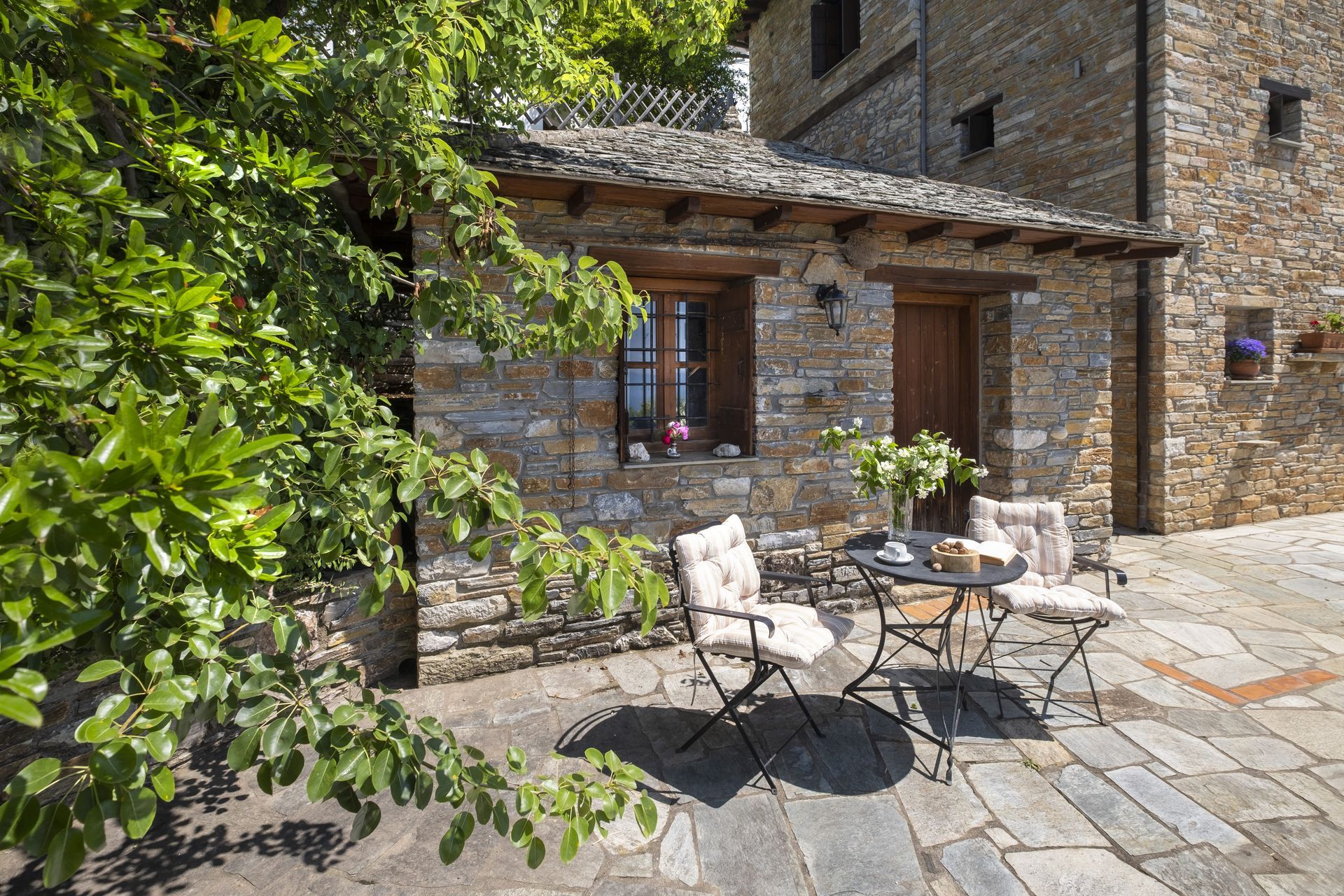Stone cottage with outdoor seating: table, two chairs, and flowers on a flagstone patio.