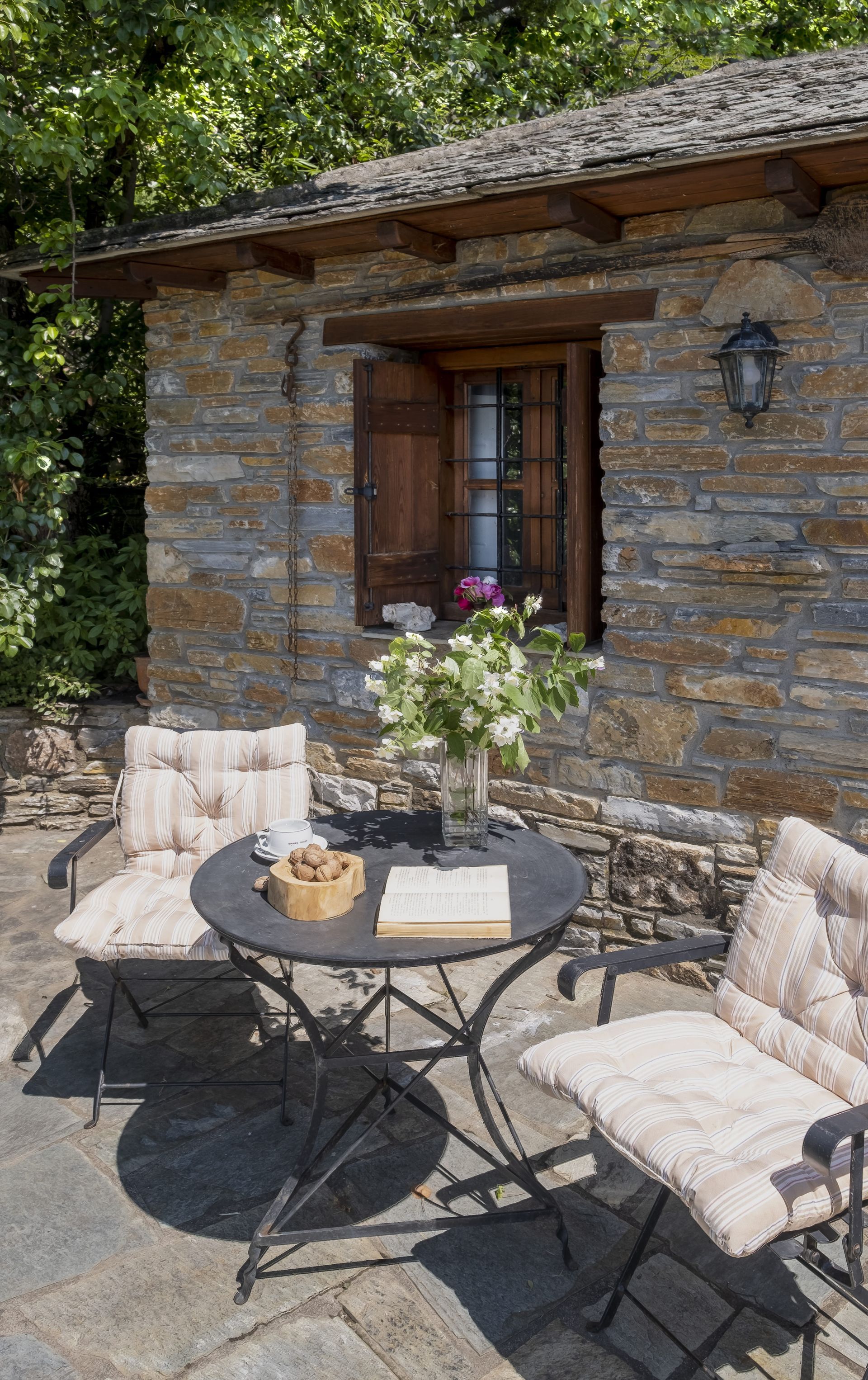 Stone cottage patio with table, chairs, flowers, and open window.