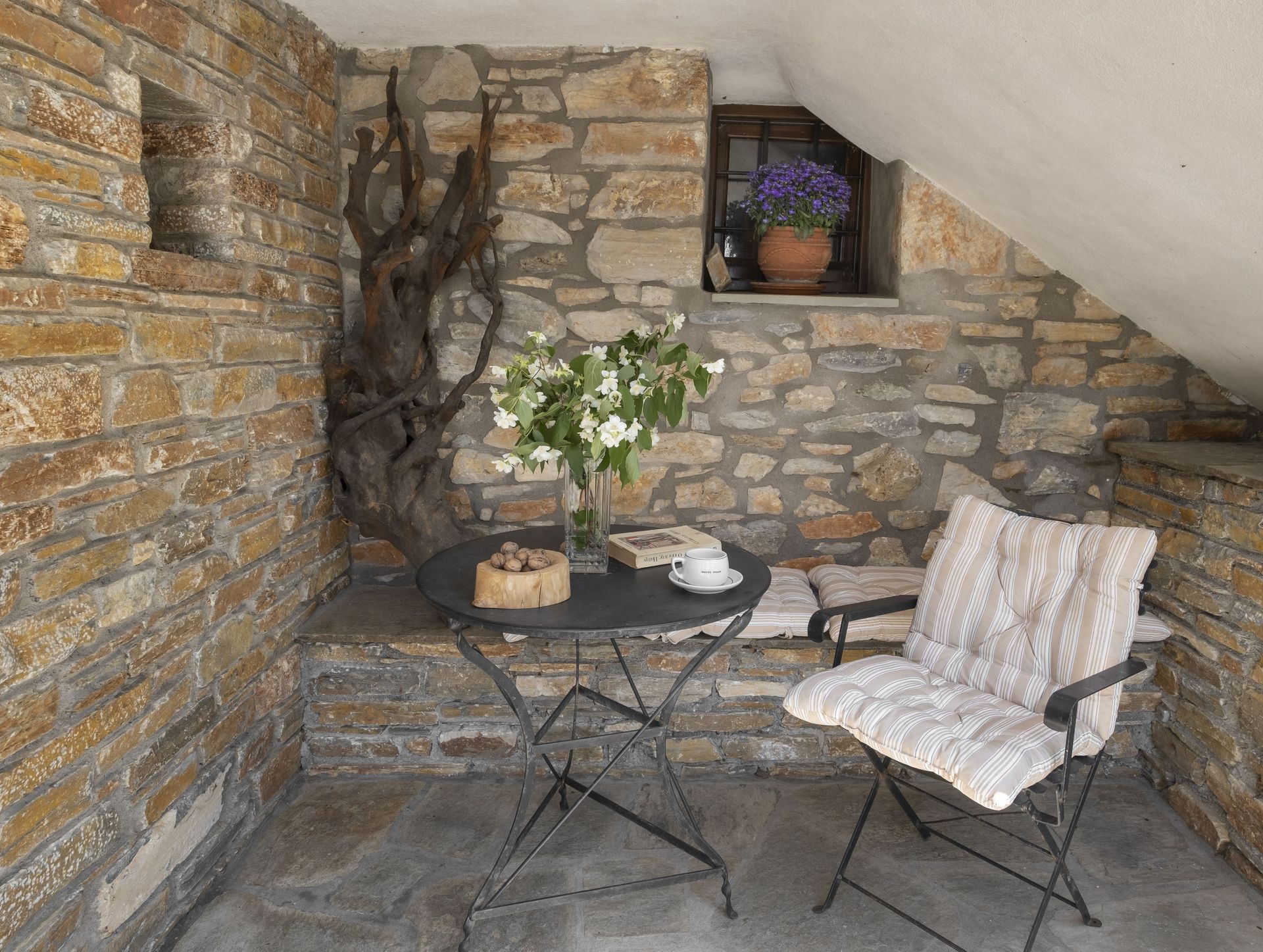 Stone-walled patio with a table, chair, flowers, and window with more flowers.