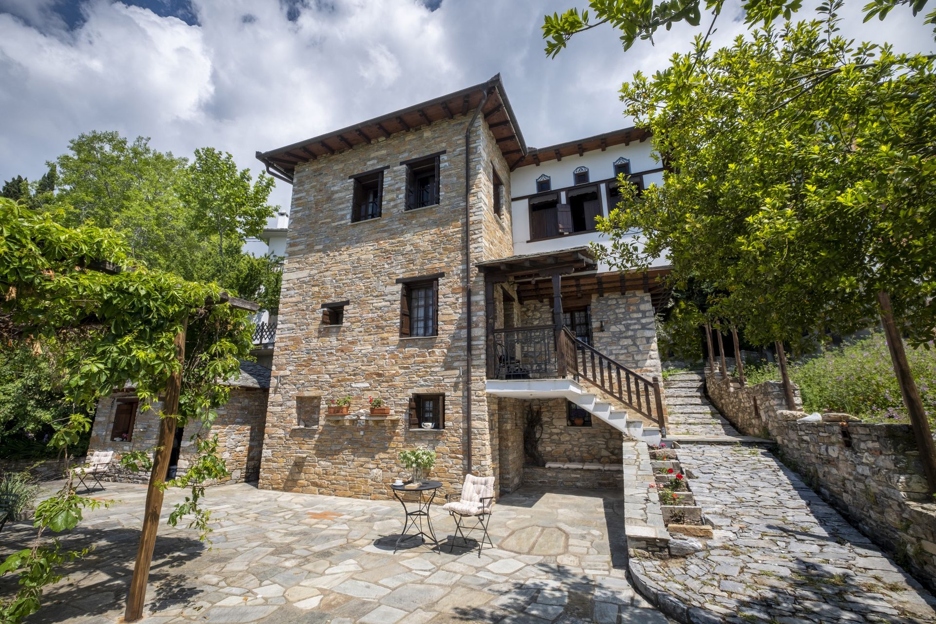 Stone house with wooden accents and stairs, nestled in a lush green setting under a cloudy sky.