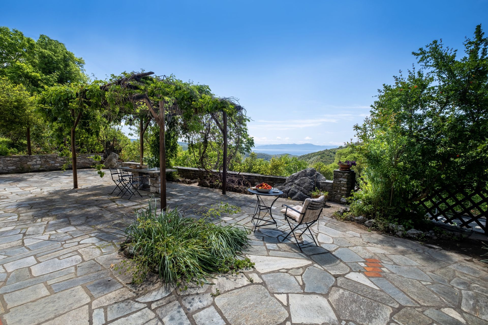 Stone patio with table and chairs under a vine-covered pergola, overlooking a distant vista.